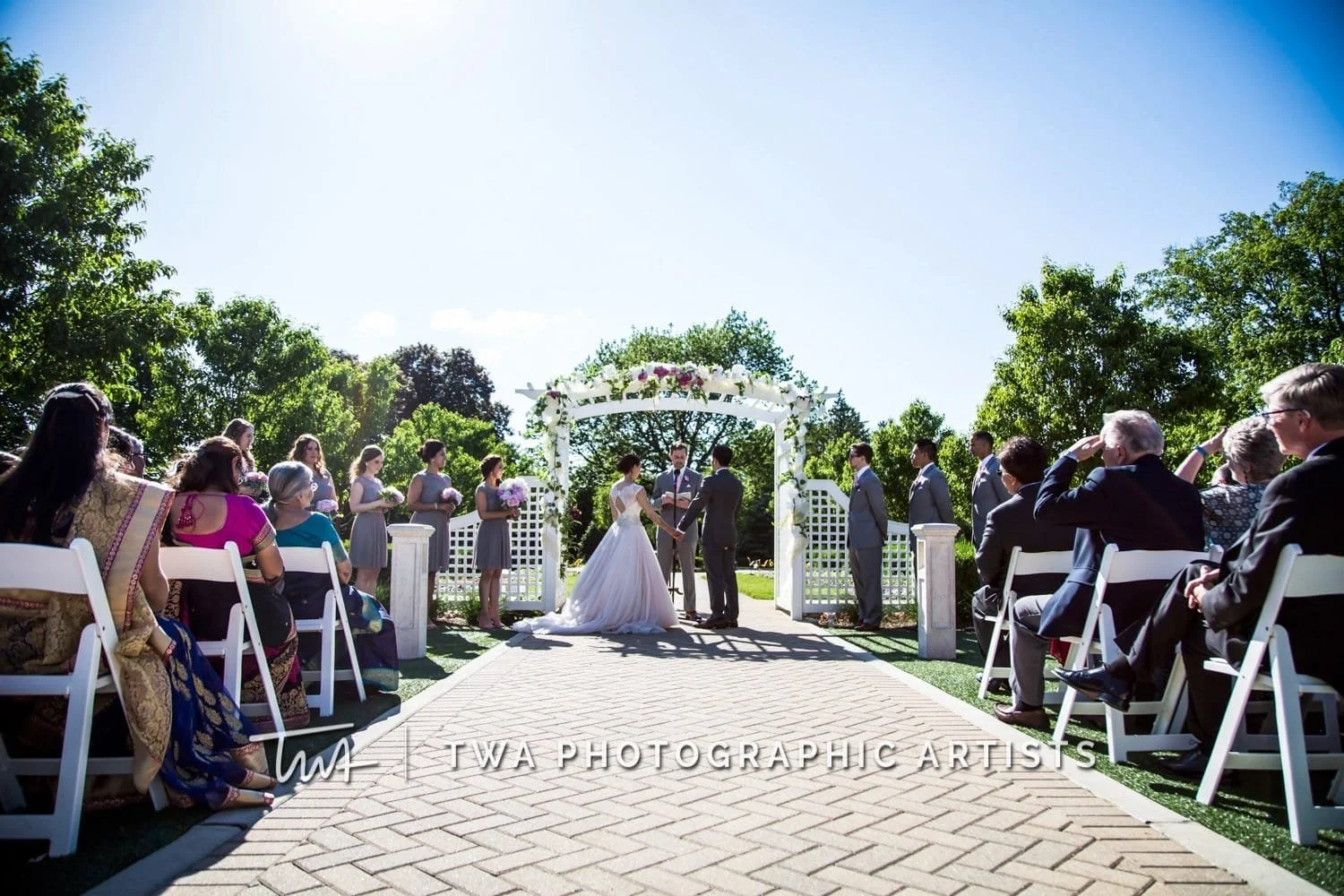 Outdoor wedding ceremony with a bride and groom holding hands under a floral arch, surrounded by seated guests and bridal party, on a sunny day with trees and blue sky in the background.