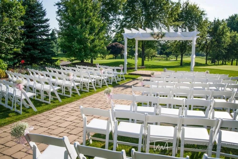 Outdoor wedding ceremony setup with white chairs, a white arbor decorated with flowers, and a brick walkway amidst green trees and grass.