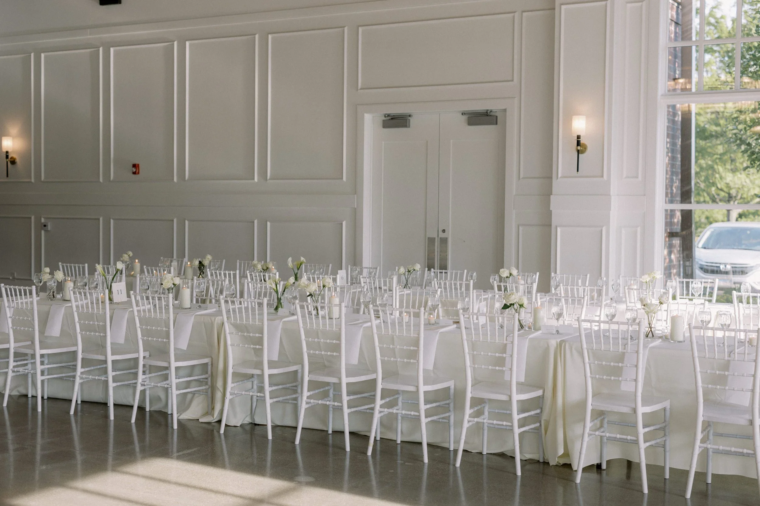 Elegant event hall decorated with white tables and chairs, with floral centerpieces and candles, large windows allowing natural light, and closed double doors in the background.