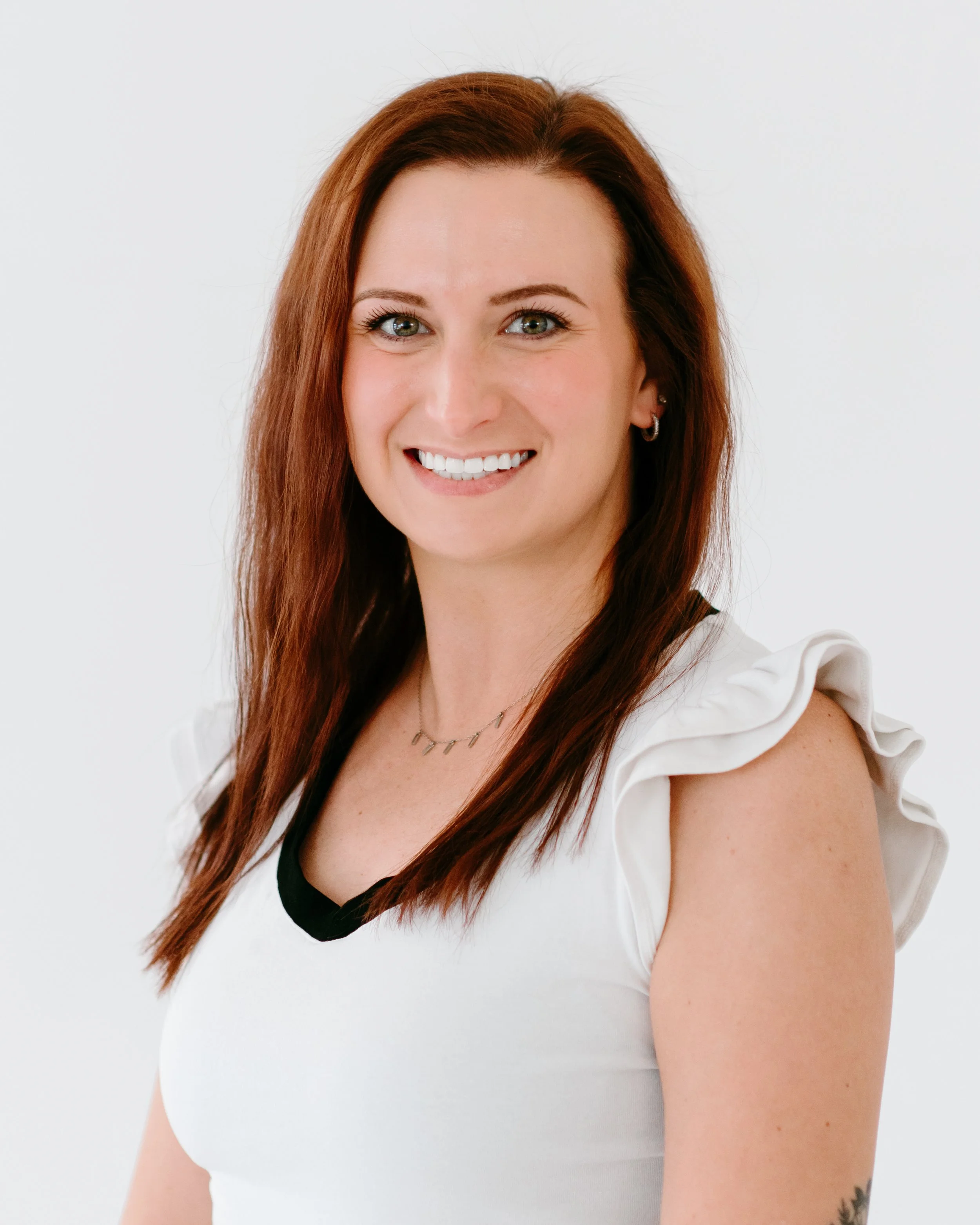 A woman with long red hair, green eyes, wearing a white top with ruffled sleeves and a black neckline, smiling against a plain white background.