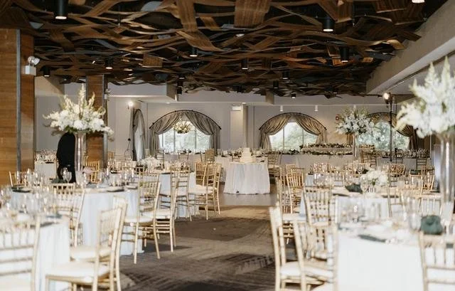 Elegant banquet hall decorated for a wedding or event with round tables, tall floral centerpieces, gold chairs, and draped windows.