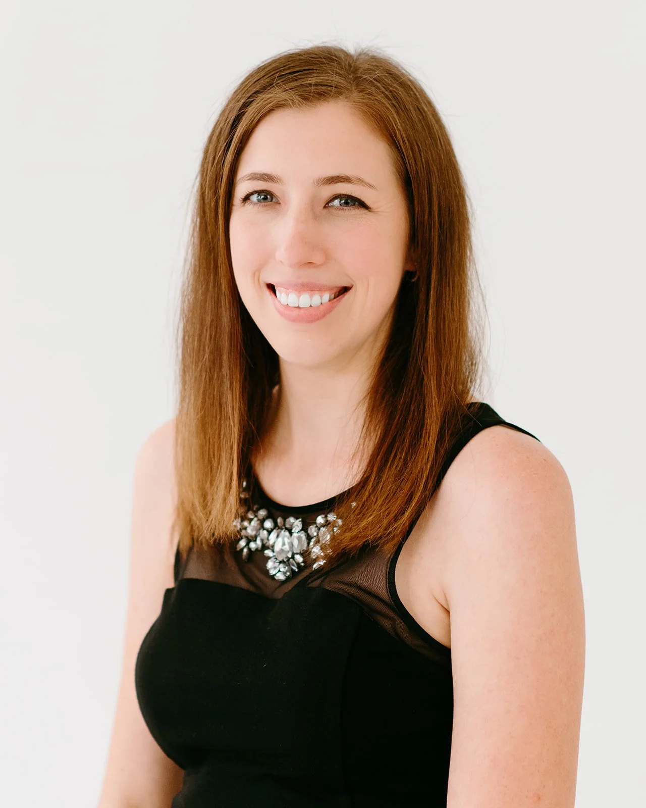A woman smiling, with long red hair, wearing a black sleeveless top with a gemstone necklace, against a white background.