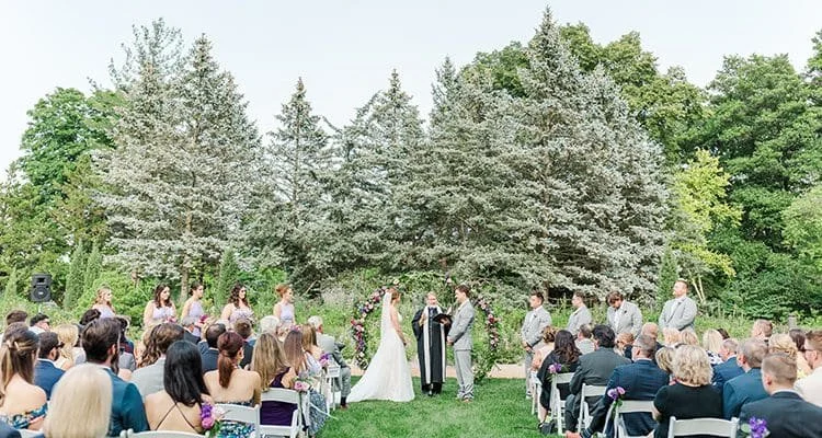 Outdoor wedding ceremony with bride and groom standing before officiant, surrounded by guests, trees, and floral decorations.
