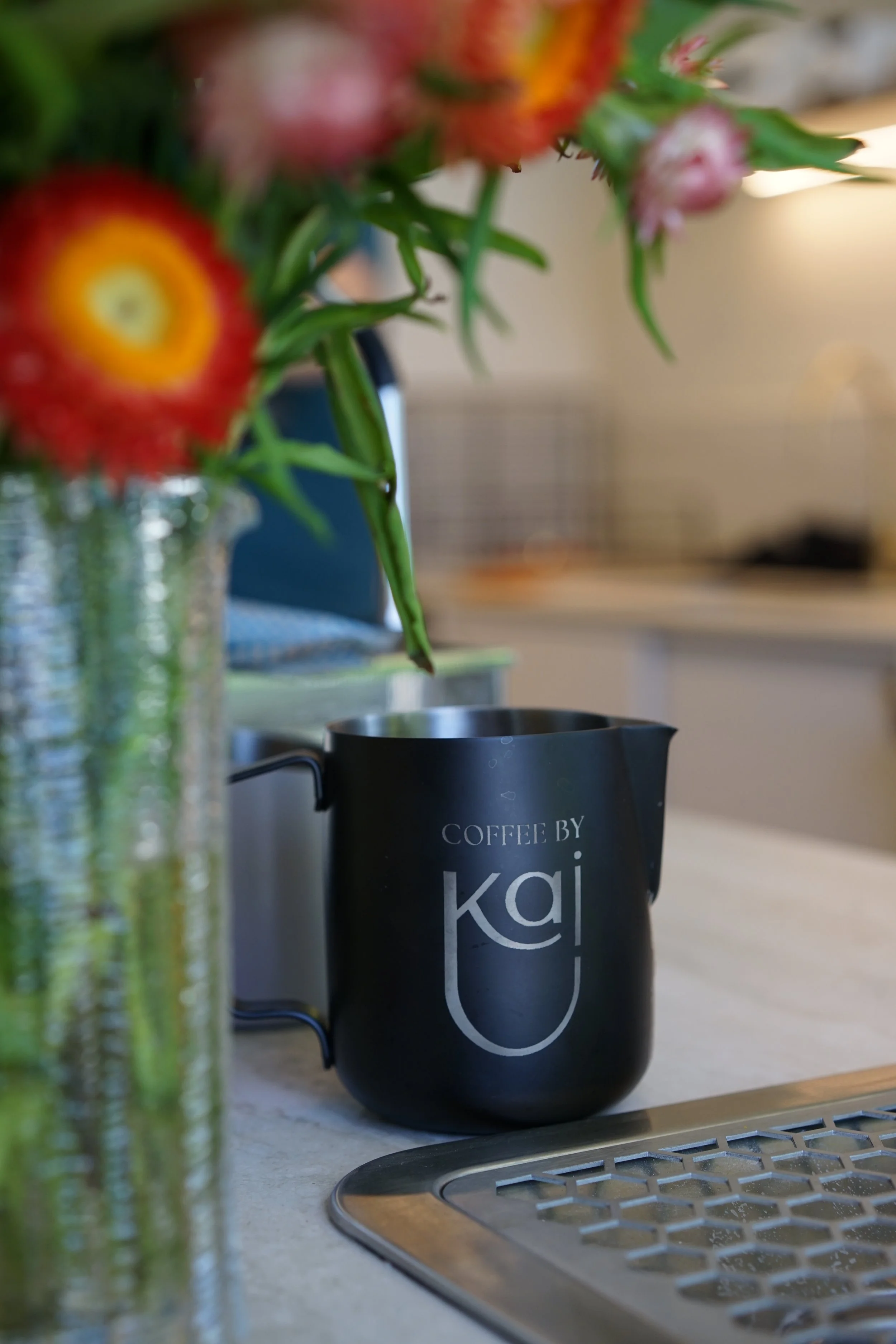 A black coloured milk container with the Coffee by Kai logo on the front of it sits on a table next to a vase of flowers.