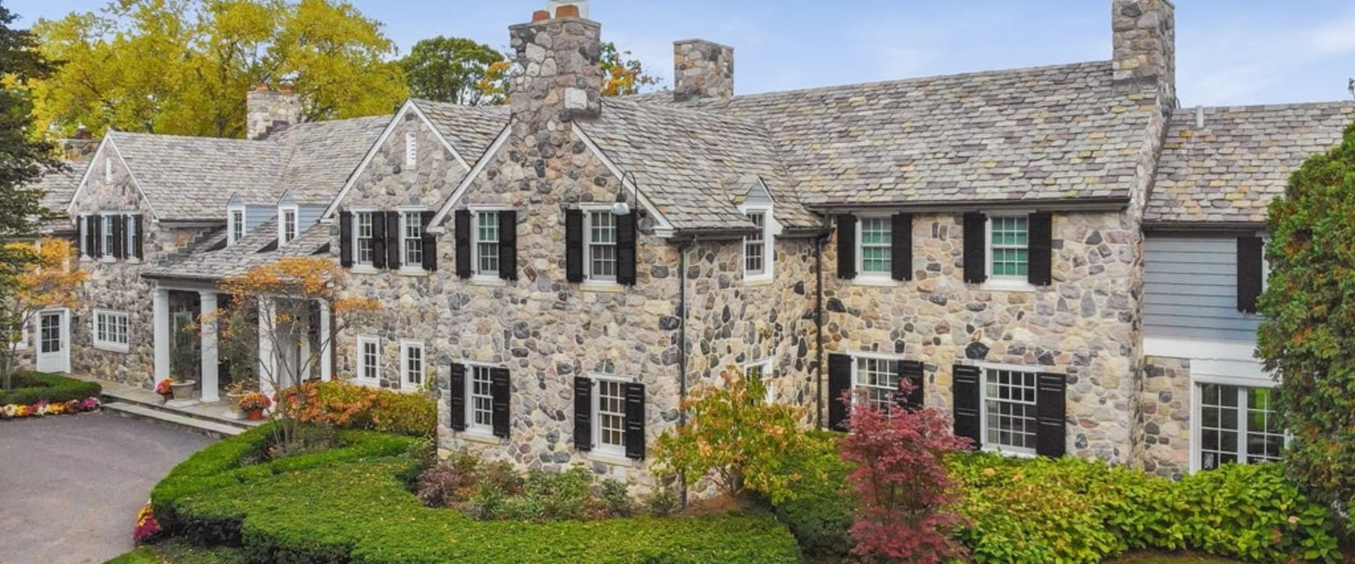 A large stone house with multiple chimneys and black shutters, surrounded by greenery and fall foliage.