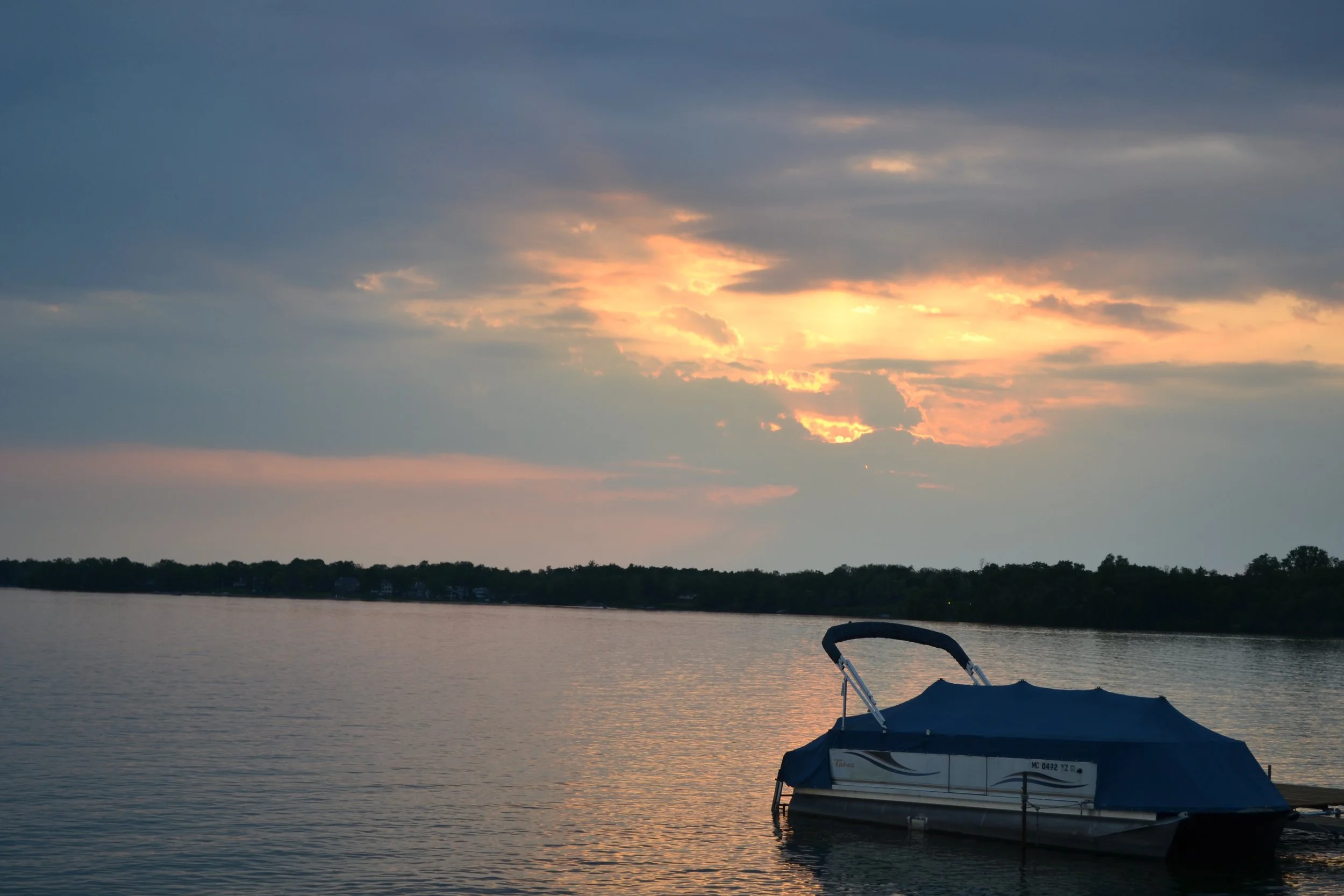 A boat covered with a blue tarp floating on calm water during sunset, with a cloudy sky and a distant shoreline in the background.