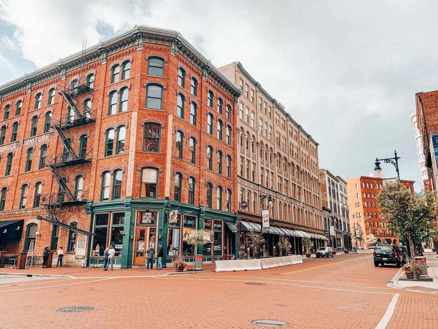 City street with brick buildings, some with fire escapes, storefronts, trees, and parked cars.