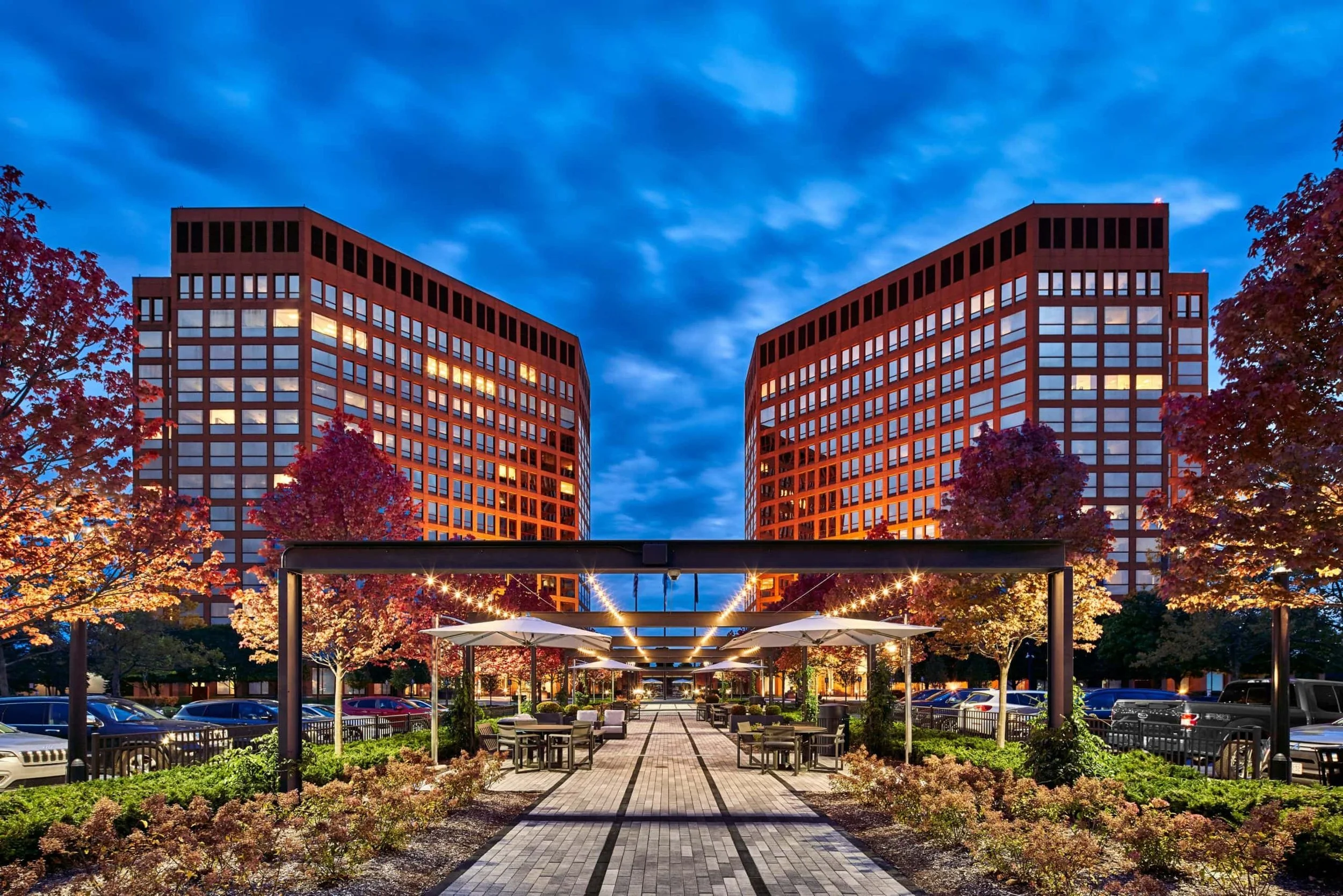 Nighttime view of modern twin office buildings with illuminated windows, surrounded by flowering trees and a landscaped outdoor seating area with tables, chairs, and string lights.
