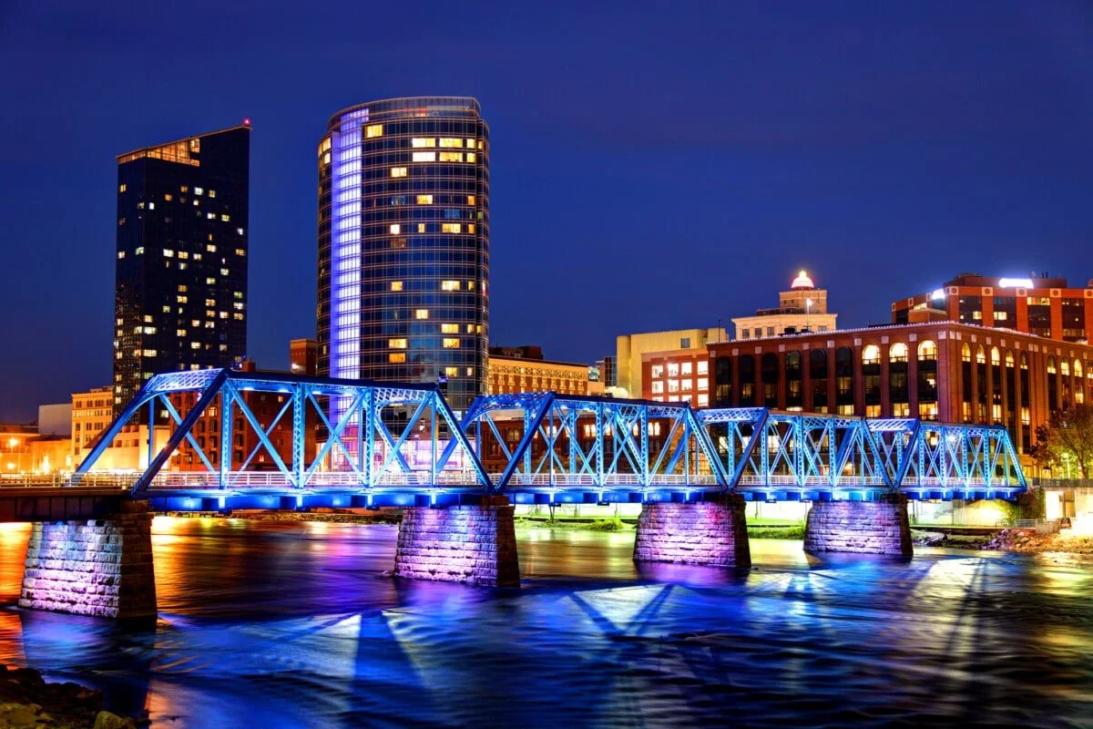 Night view of a city skyline with illuminated buildings and a blue lit bridge over a body of water.