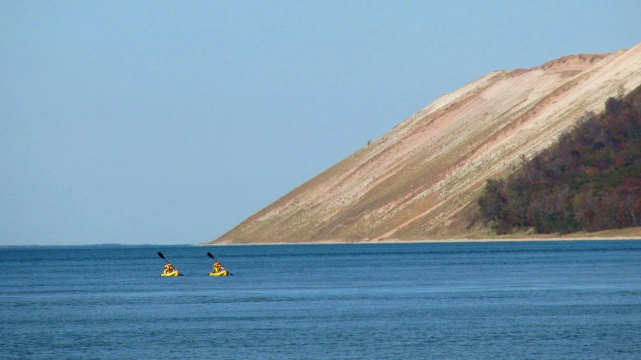 Two people kayaking in a large body of water with a sloped hill in the background.