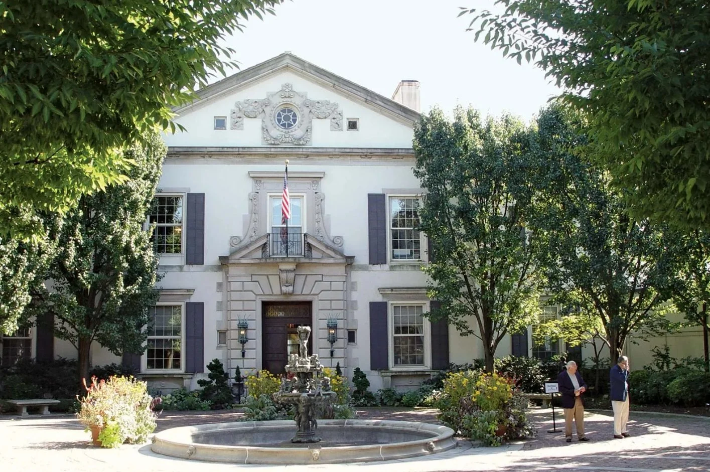 Two men in suits standing and talking in front of a historic white building with windows and an American flag, surrounded by trees and garden with a fountain in the foreground.