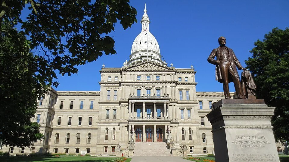 The state capitol building with a large white dome, surrounded by trees, and a bronze statue of Austin Blair, labeled as War Governor of Michigan, in the foreground.