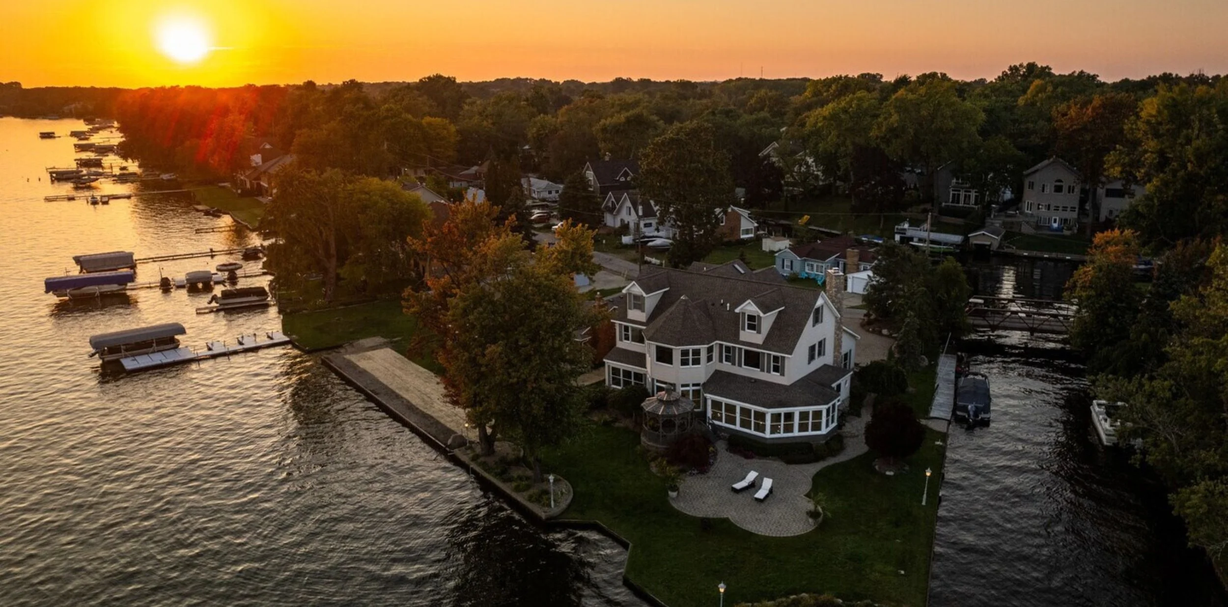 An aerial view of a lakeside house at sunset with a large yard, boat docks, multiple boats, and a waterway lined with trees and neighboring houses.