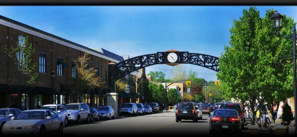 A busy downtown street with parked cars along the sidewalk, a decorative arch with a clock overhead, trees lining the street, people walking on the sidewalk, and a clear blue sky.