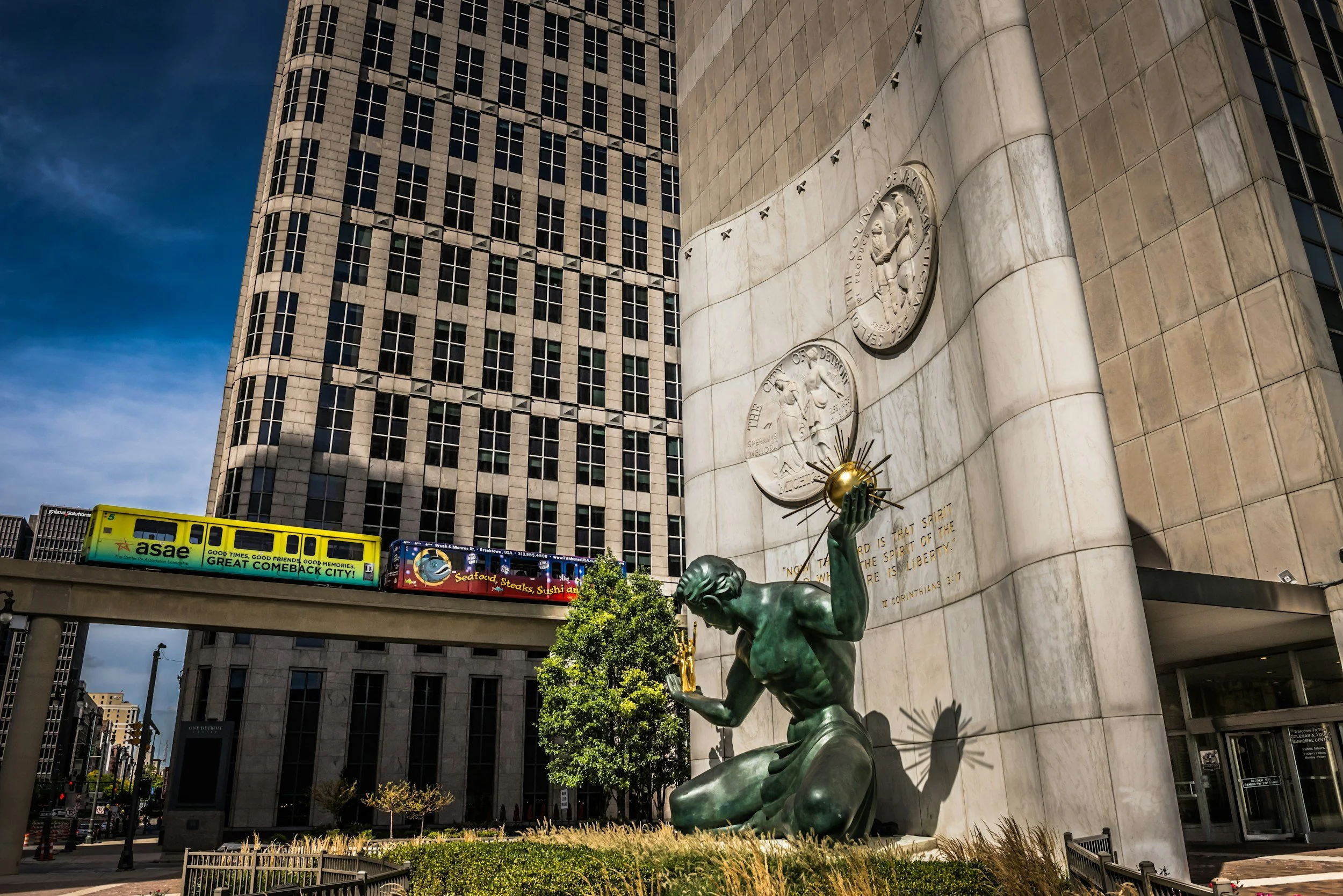A large modern building with a stone facade, featuring a bronze kneeling sculpture in front. The sculpture depicts a figure holding a globe with rays emanating from it, placed at the corner of the building. An elevated train track with colorful adver