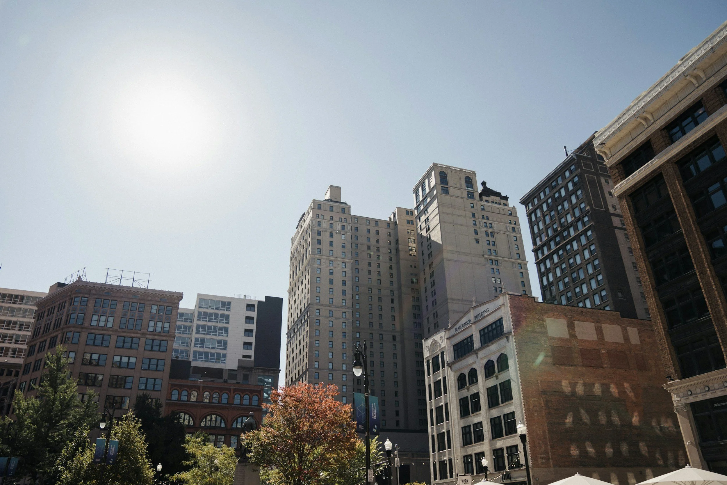 Downtown cityscape with tall buildings, trees, and outdoor seating area under a bright sun.