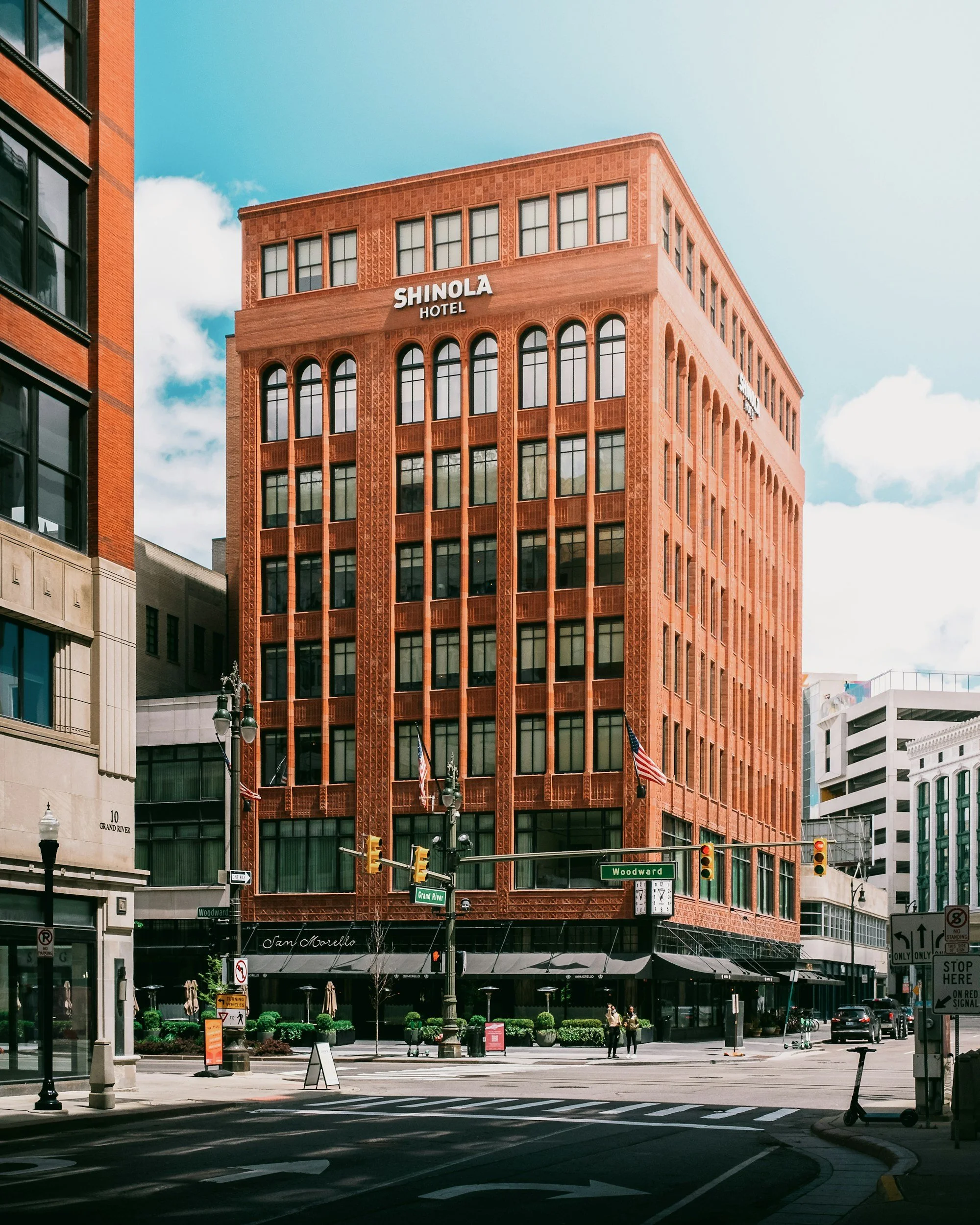 A tall red brick hotel building with the sign 'Shinola Hotel' at the top, located at the corner of Woodward Avenue and Grand River in an urban cityscape with a few pedestrians and cars.