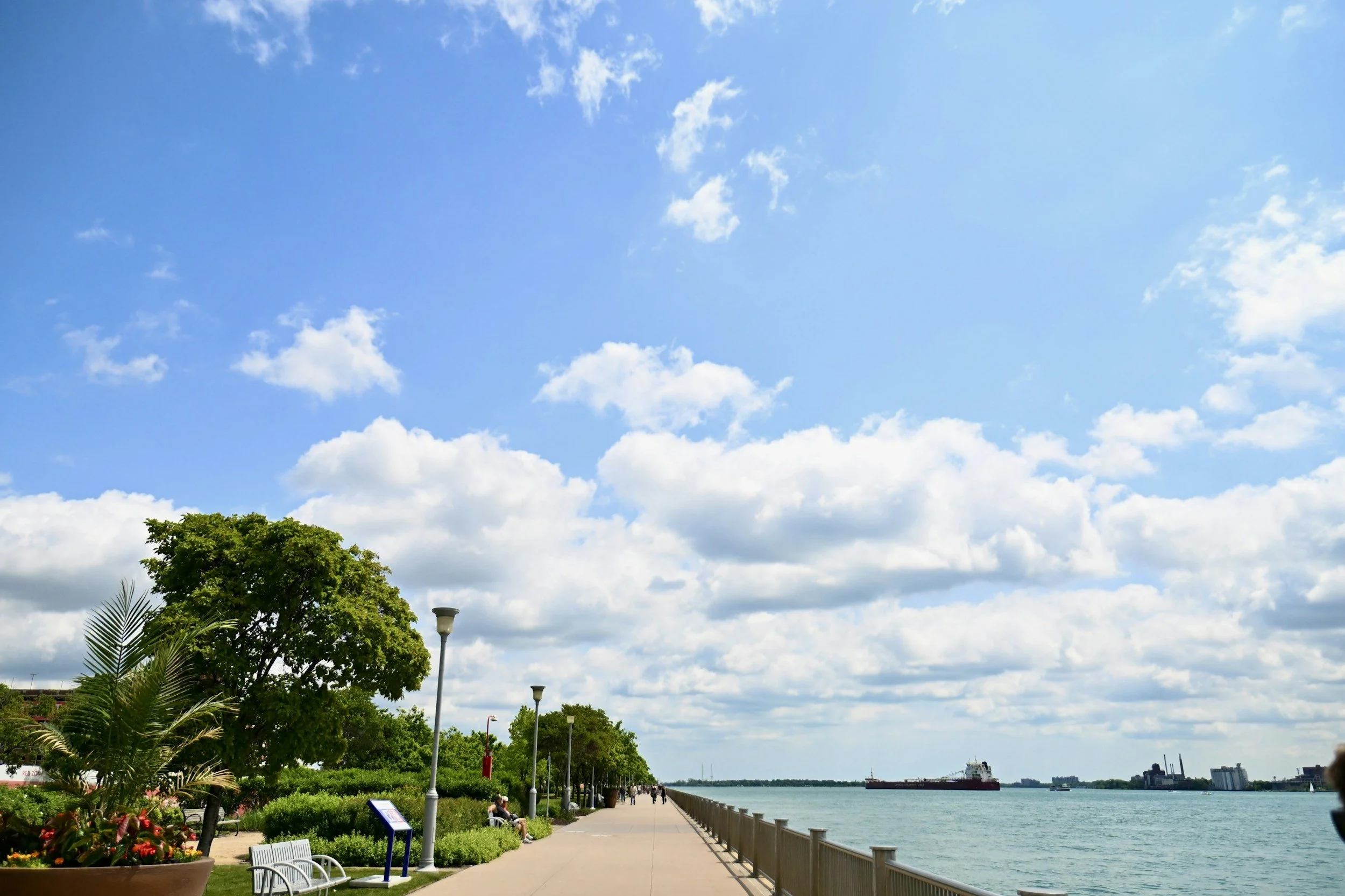 A waterfront park with a paved walking path, benches, street lamps, trees, bushes, and flowers, overlooking a body of water with ships and a city skyline in the distance, under a partly cloudy sky.