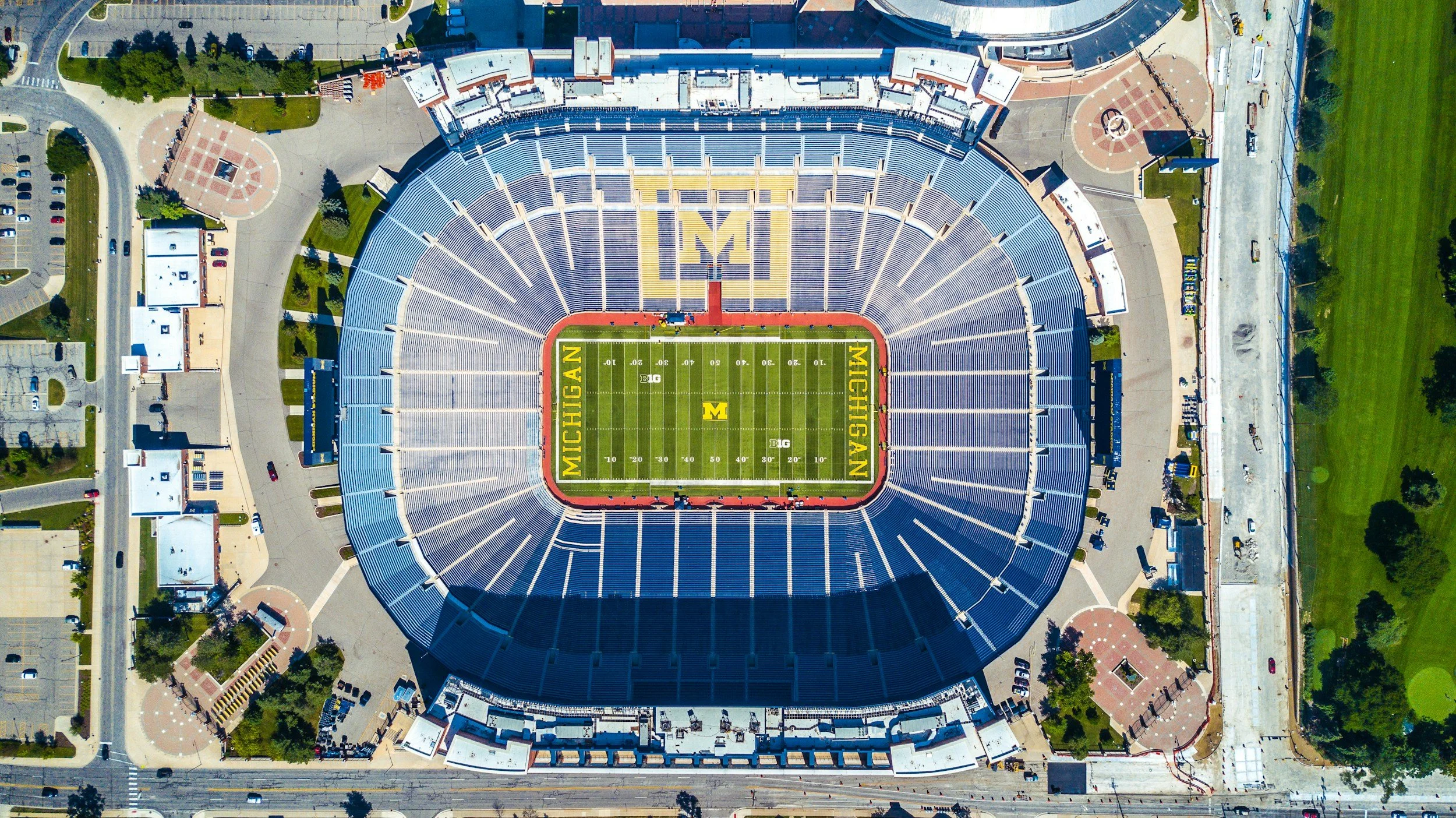 Aerial view of Michigan Stadium, also known as the Big House, with a football field in the center, marked with Michigan and Big Ten logos, surrounded by empty blue seats, walkways, and parking areas.