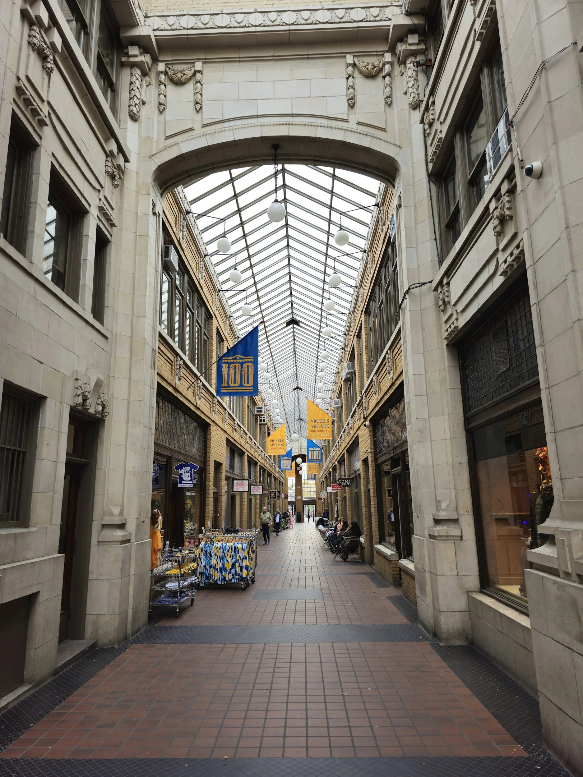 Indoor shopping arcade with a glass roof, banners reading '100' and 'Nickels Arcade,' stores on both sides, and people walking and sitting.