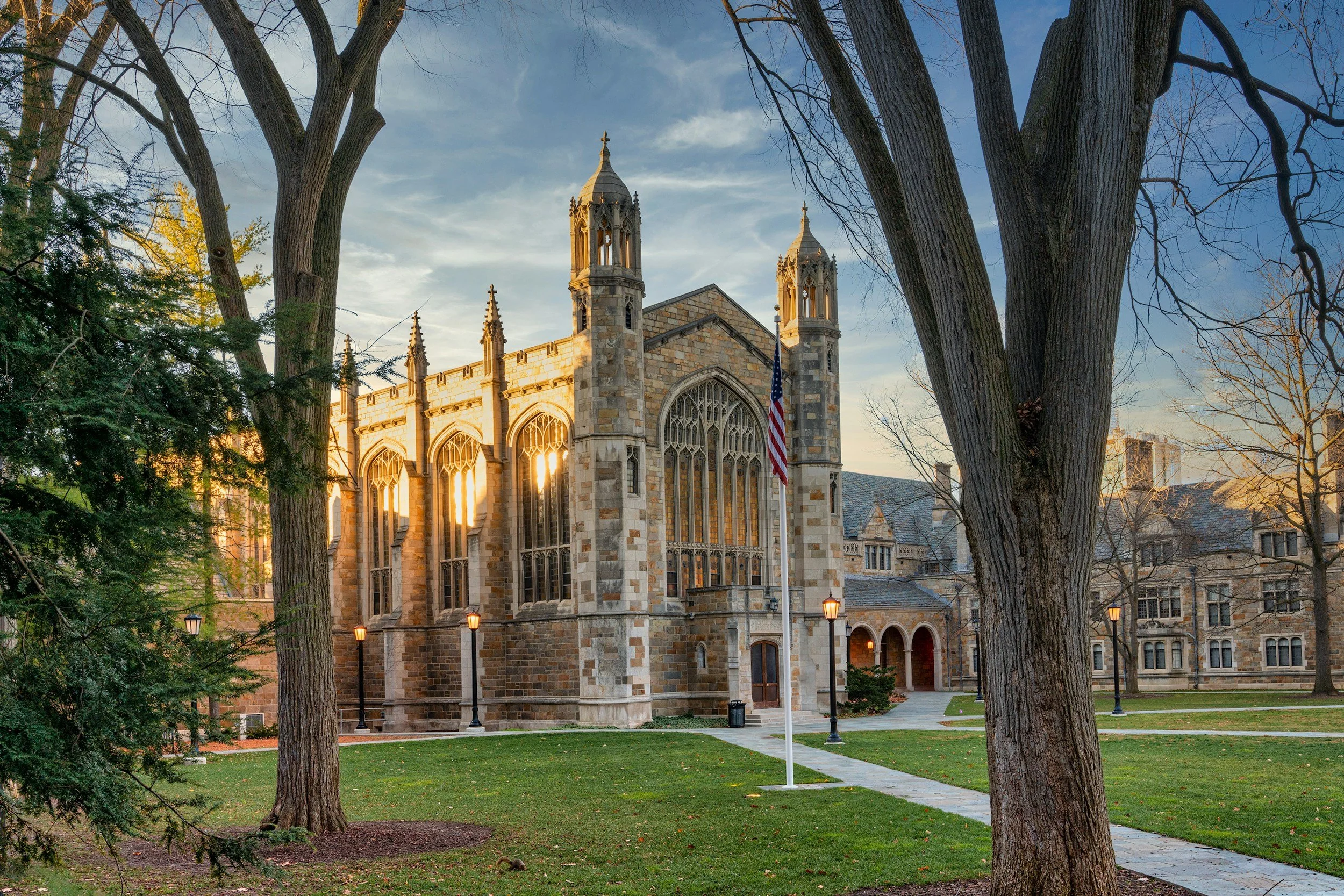 A historic stone church with tall windows and gothic architecture, surrounded by trees and illuminated by streetlights at sunset.