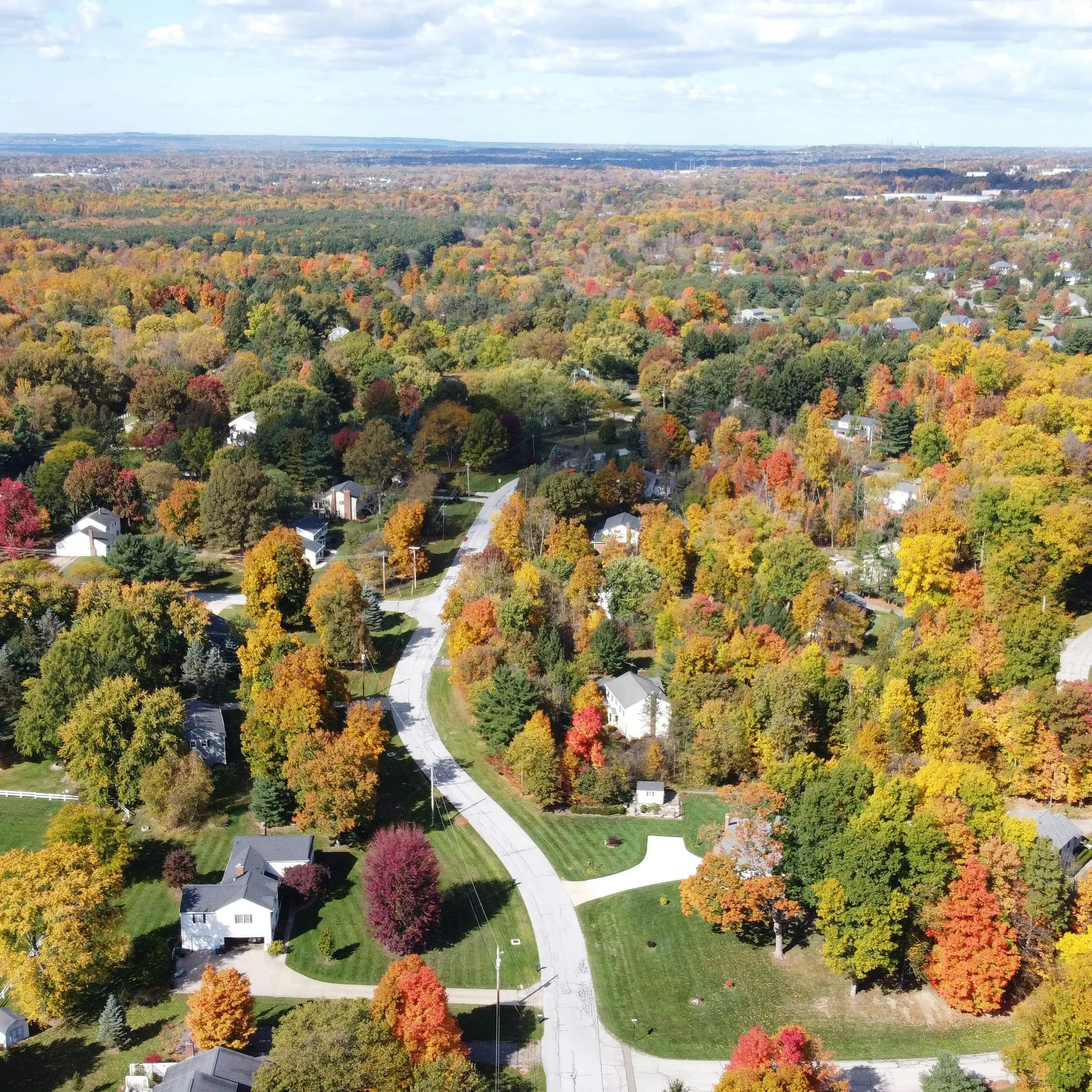 Aerial view of a suburban neighborhood during fall, showing colorful autumn trees, winding roads, and houses with well-maintained yards.