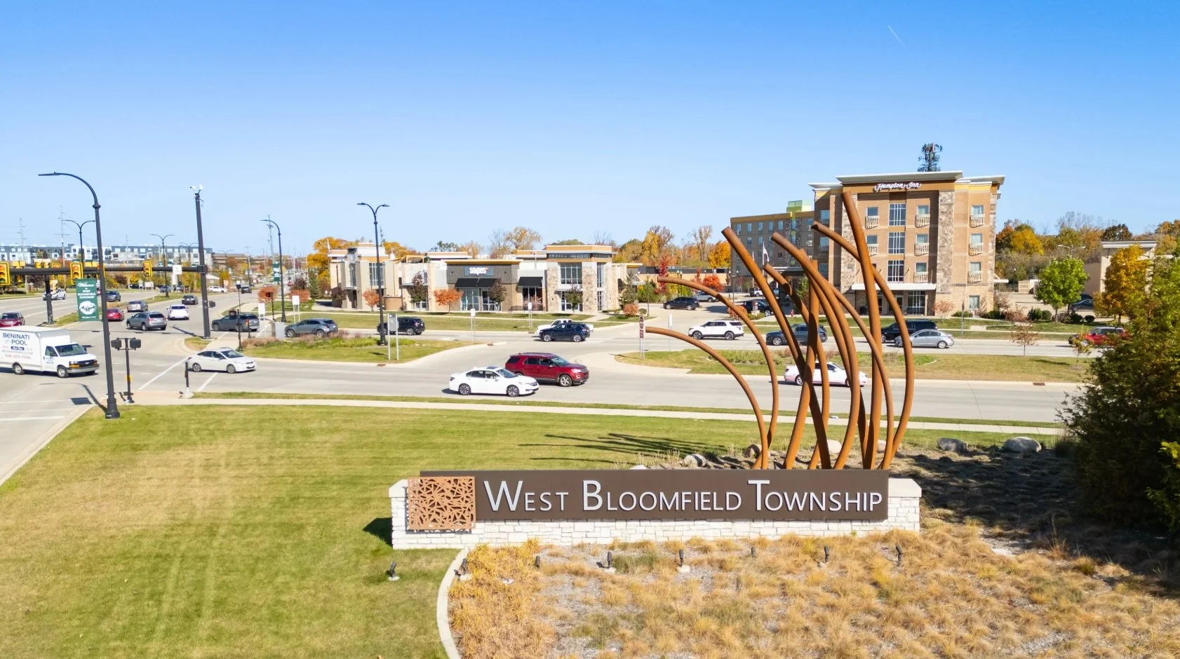 Sign for West Bloomfield Township with decorative sculpture, across parking lot and buildings, trees with fall foliage in background, clear blue sky.