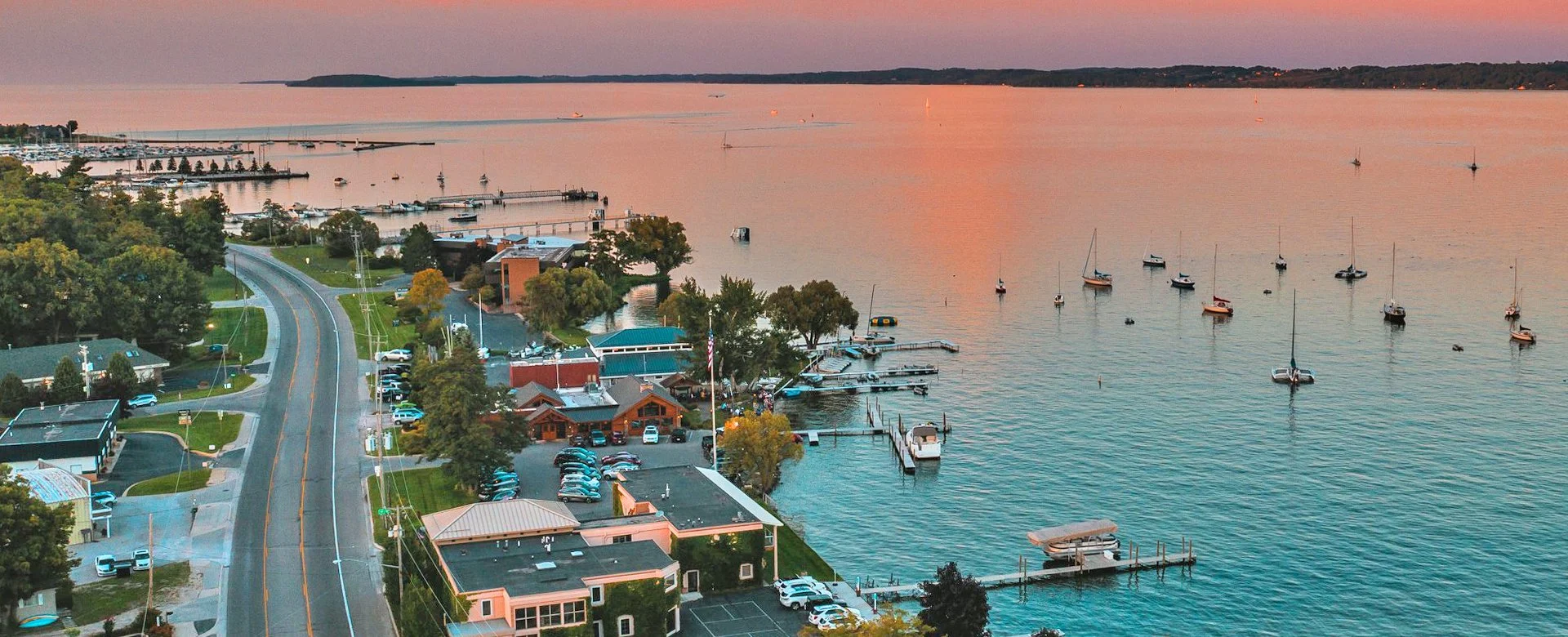 Aerial view of a harbor during sunset with sailboats anchored in calm water, a shoreline with buildings and parking lot, and a curved road lined with trees.
