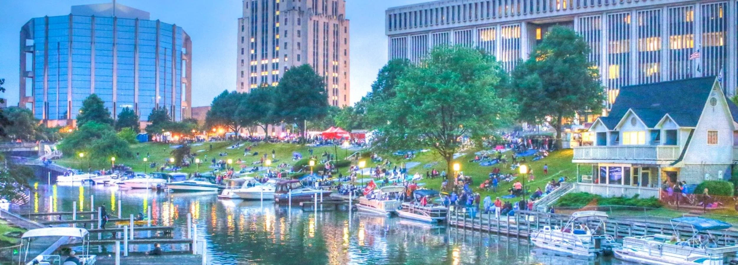 A lively waterfront scene in a city park during the evening, featuring boats docked along the water, a grassy area with many people relaxing and socializing, and tall modern buildings in the background.