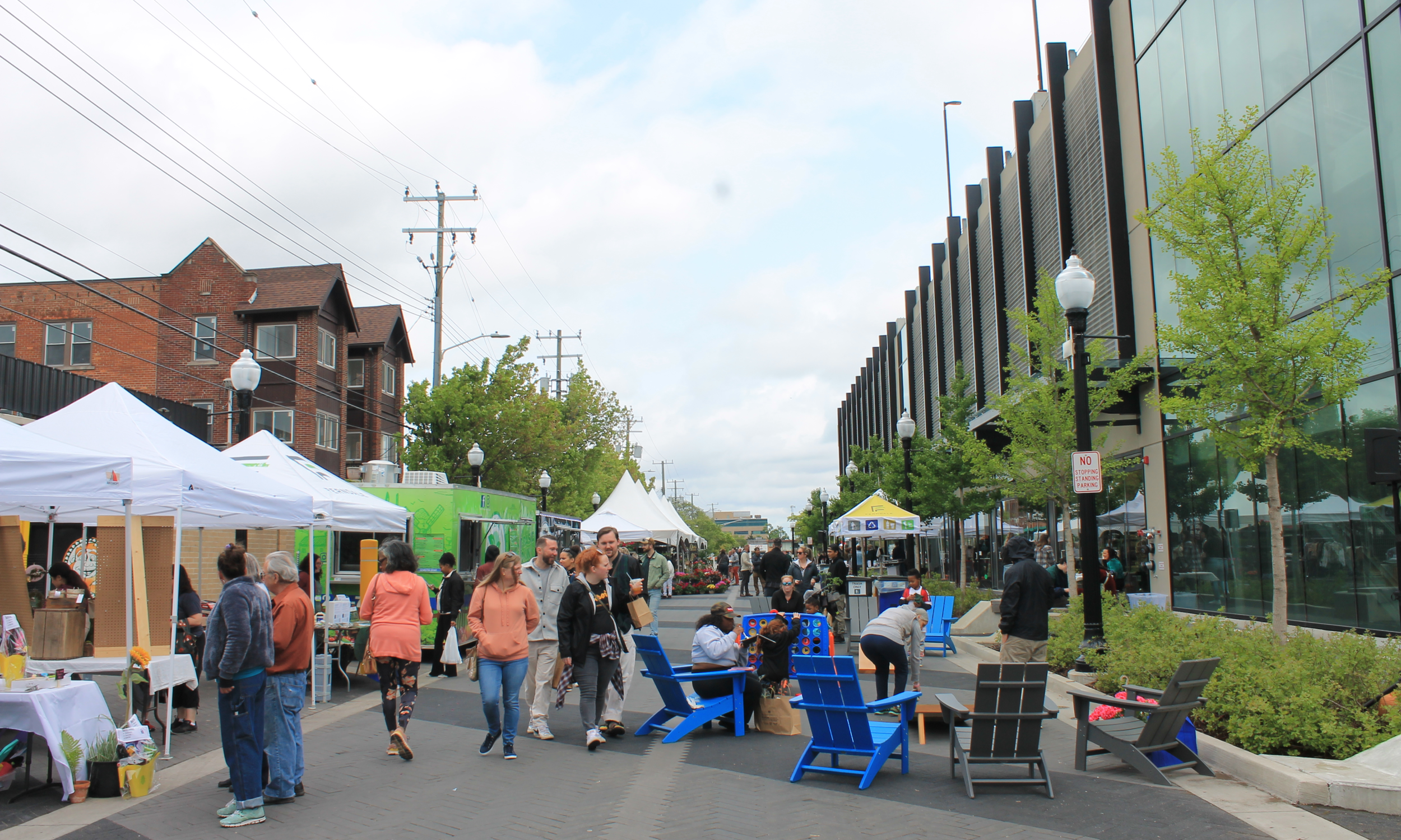 People walking and sitting at an outdoor market with tents, food trucks, benches, and trees on a paved street, with a modern glass building on the right and brick buildings on the left.