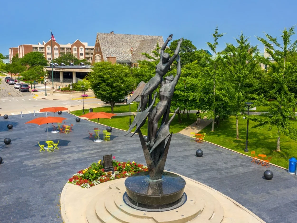 Public plaza with a bronze sculpture of three abstract human figures reaching upward, surrounded by colorful tables and umbrellas, trees, and a parking lot in the background.