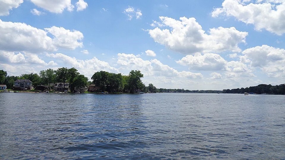 Calm river with houses and trees along the shoreline, and a partly cloudy sky.
