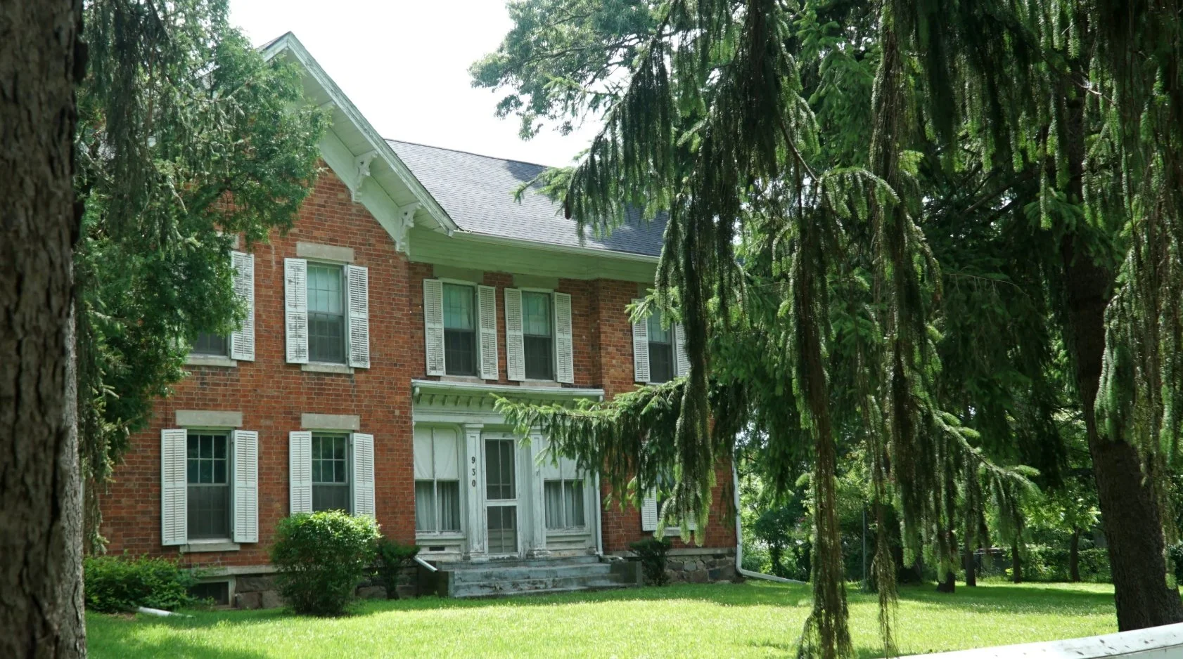 A two-story brick house with white shutters and steps leading to the front door, surrounded by green trees and a well-maintained lawn.