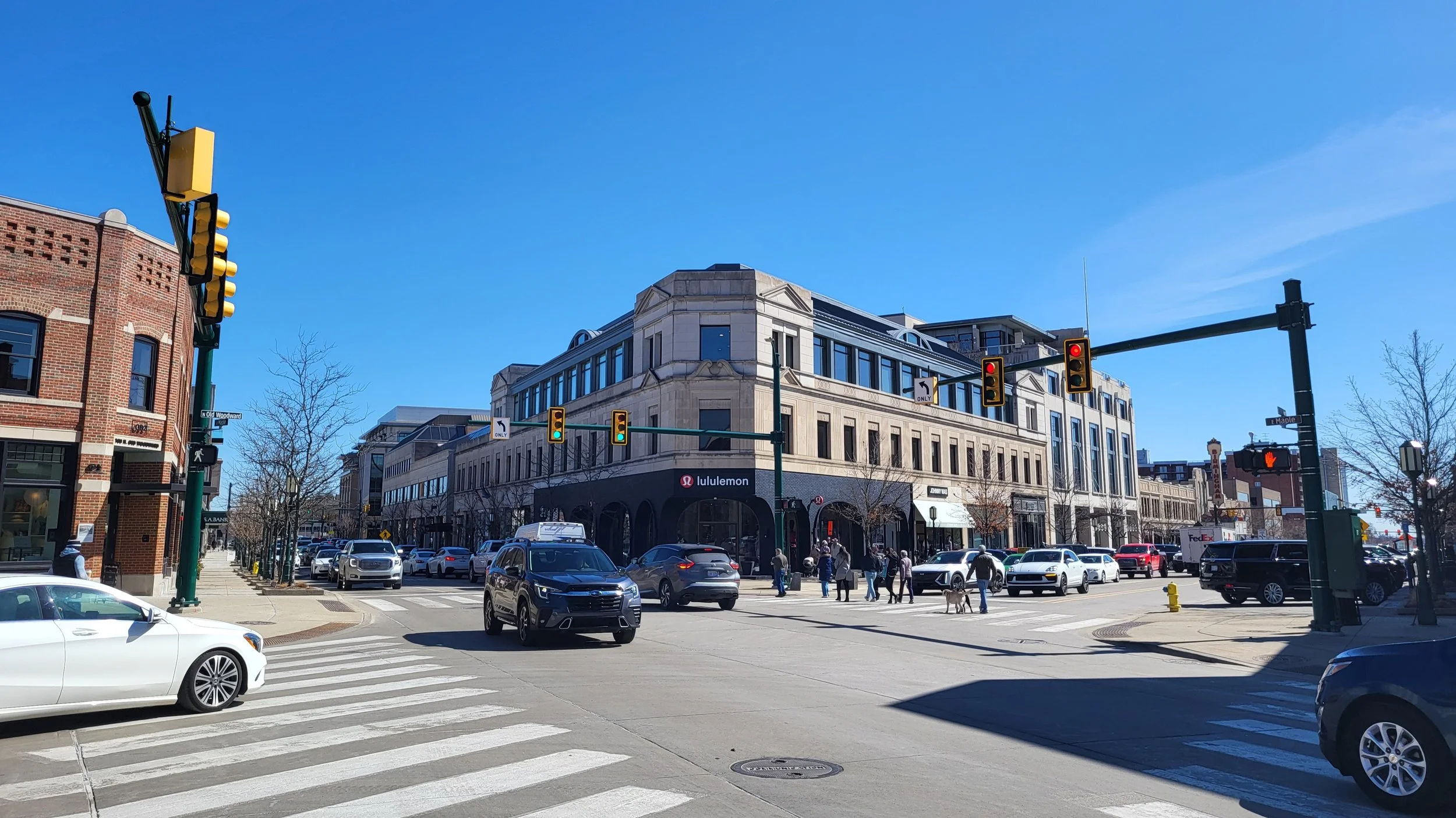 City intersection with crosswalks, cars, pedestrians, traffic lights, and buildings including a Lululemon store on a sunny day.