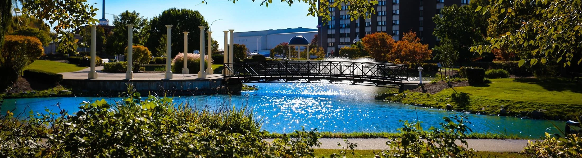 A park with a small bridge over a pond, surrounded by trees and bushes with colorful leaves, and an apartment building and a large industrial building in the background, on a sunny day.