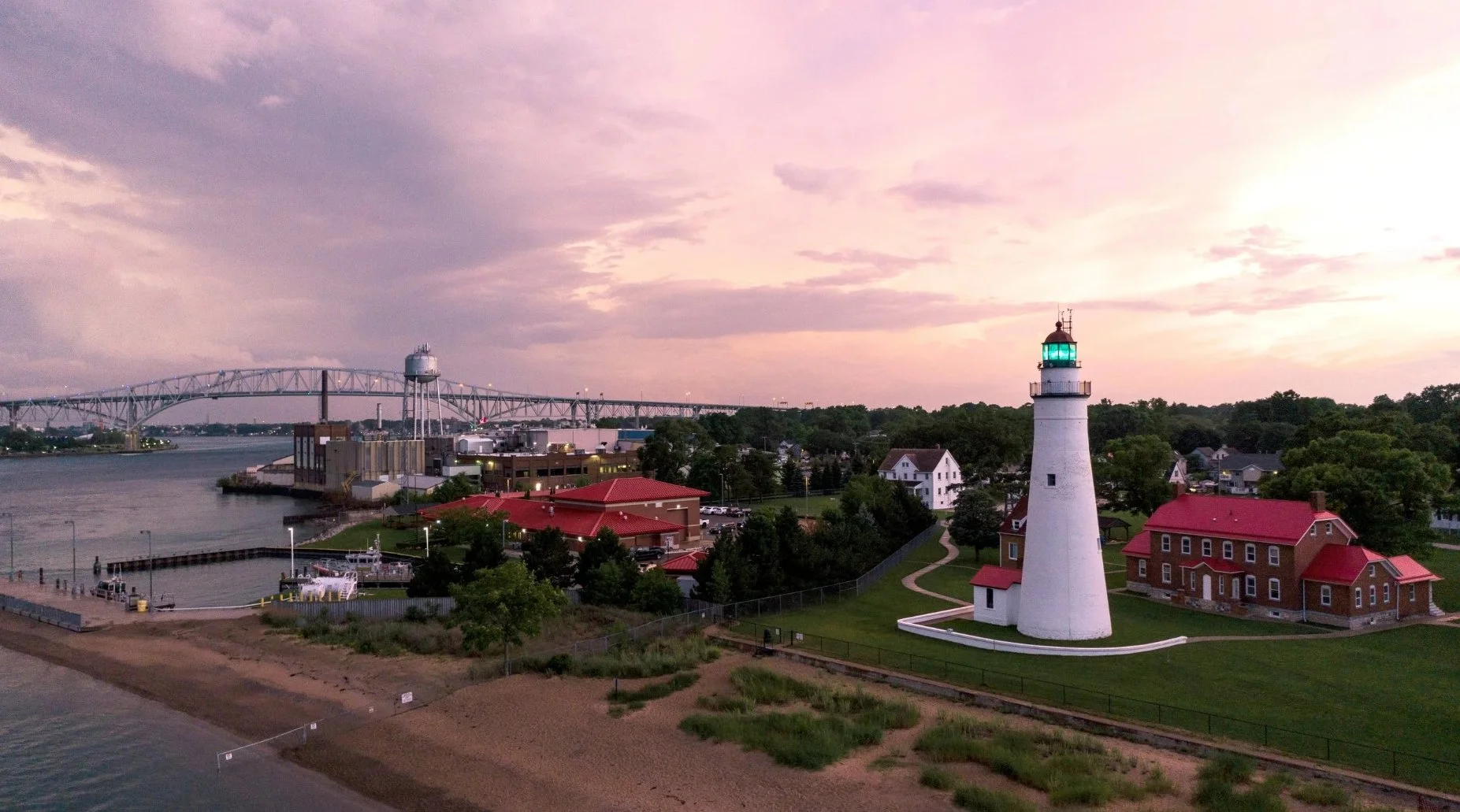 A lighthouse with a black top and green light shining, situated on a grassy area near a beach, with houses and trees in the background, under a pink and purple sky at sunset.