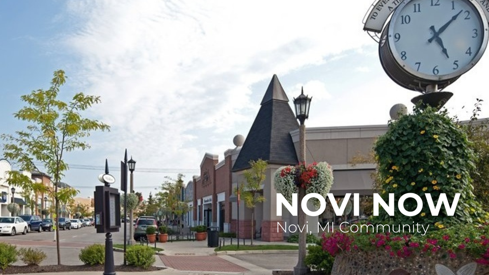 Downtown street scene in Novi, Michigan with decorative clock, lush flower arrangements, storefronts, trees, and parked cars under a partly cloudy sky.
