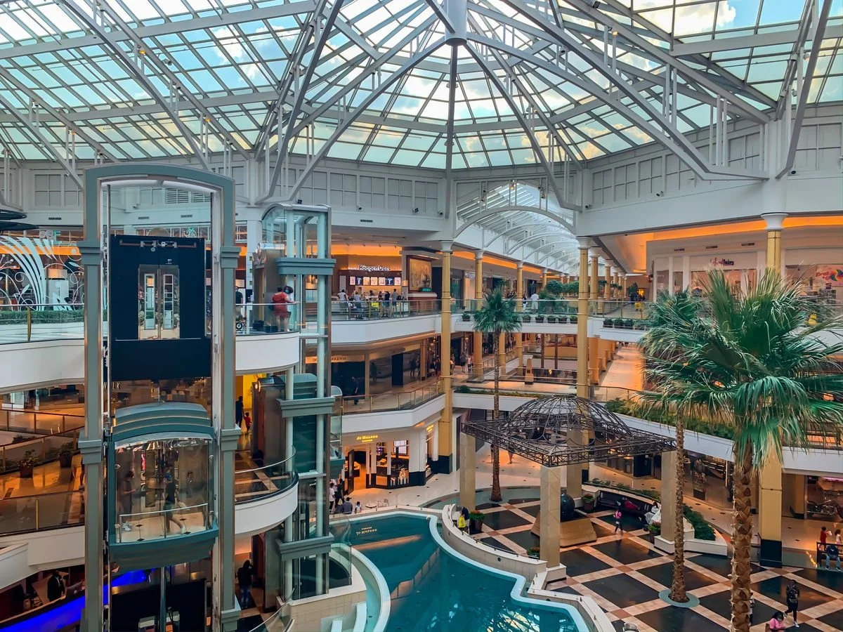 Interior view of a multi-level shopping mall with a glass ceiling, escalators, palm trees, and several stores and shoppers.