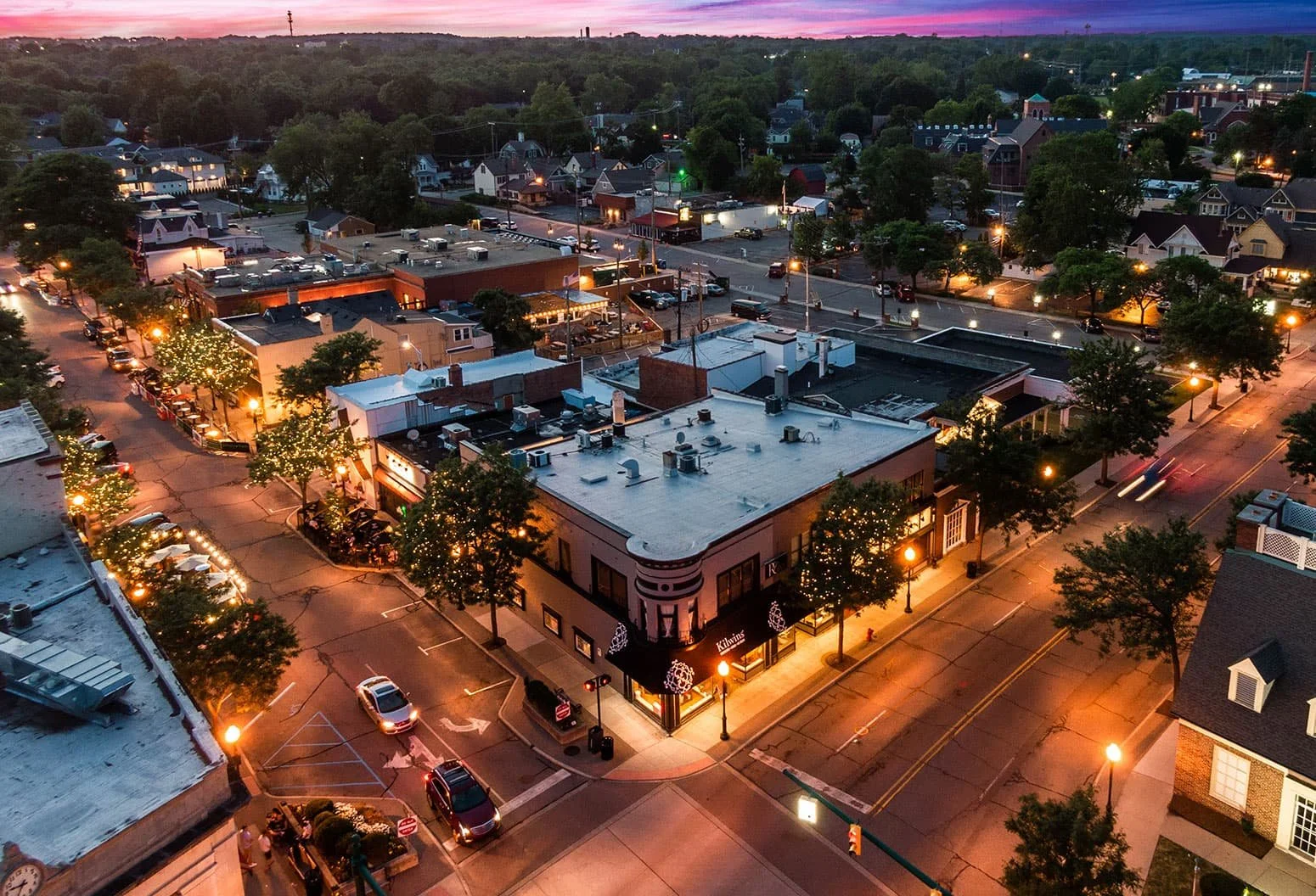 Aerial view of downtown main street during dusk, with buildings decorated with holiday lights and trees, and cars parked along the streets.