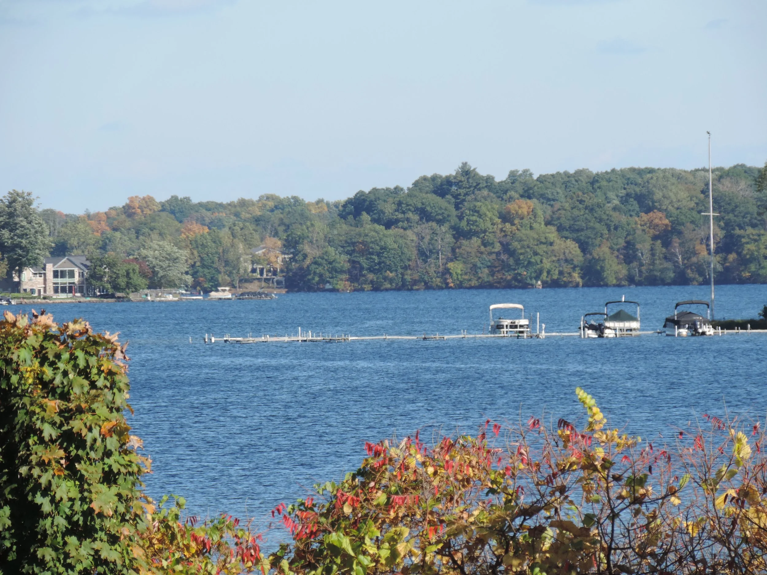 A peaceful lakeside scene with boats docked at a pier, surrounded by trees with fall foliage and residential houses in the background.