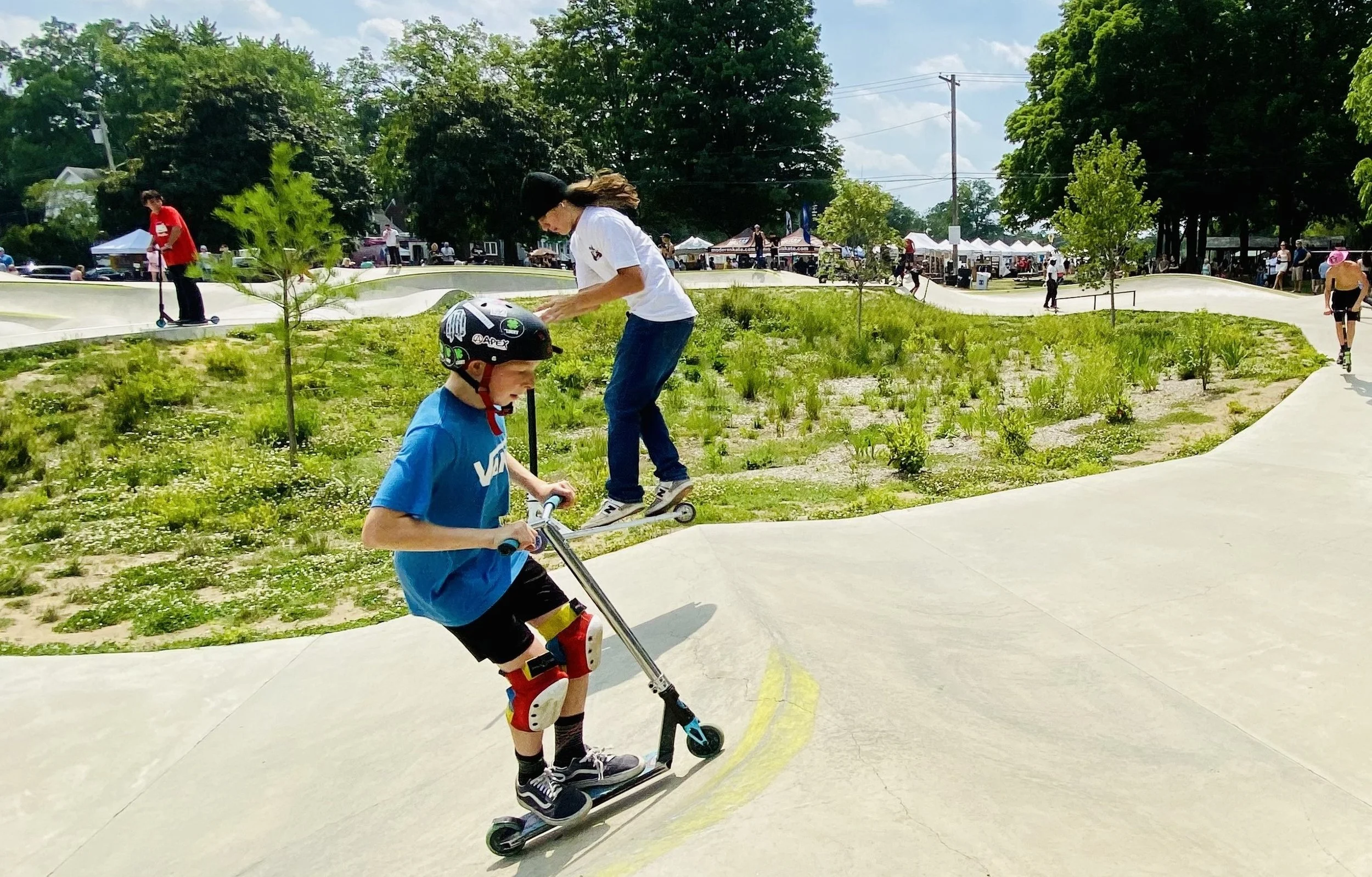 Two children riding scooters on a skatepark with green grass, trees, and people in the background.