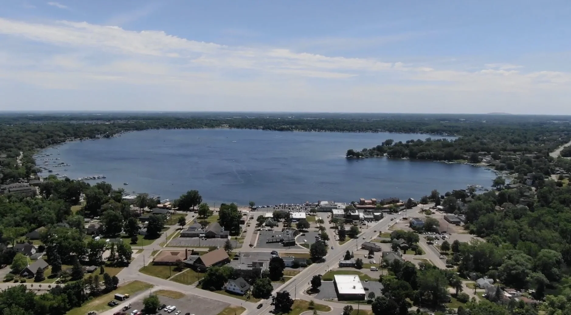 Aerial view of a town around a large, calm lake with boats and docks, surrounded by green trees and residential houses under a partly cloudy blue sky.