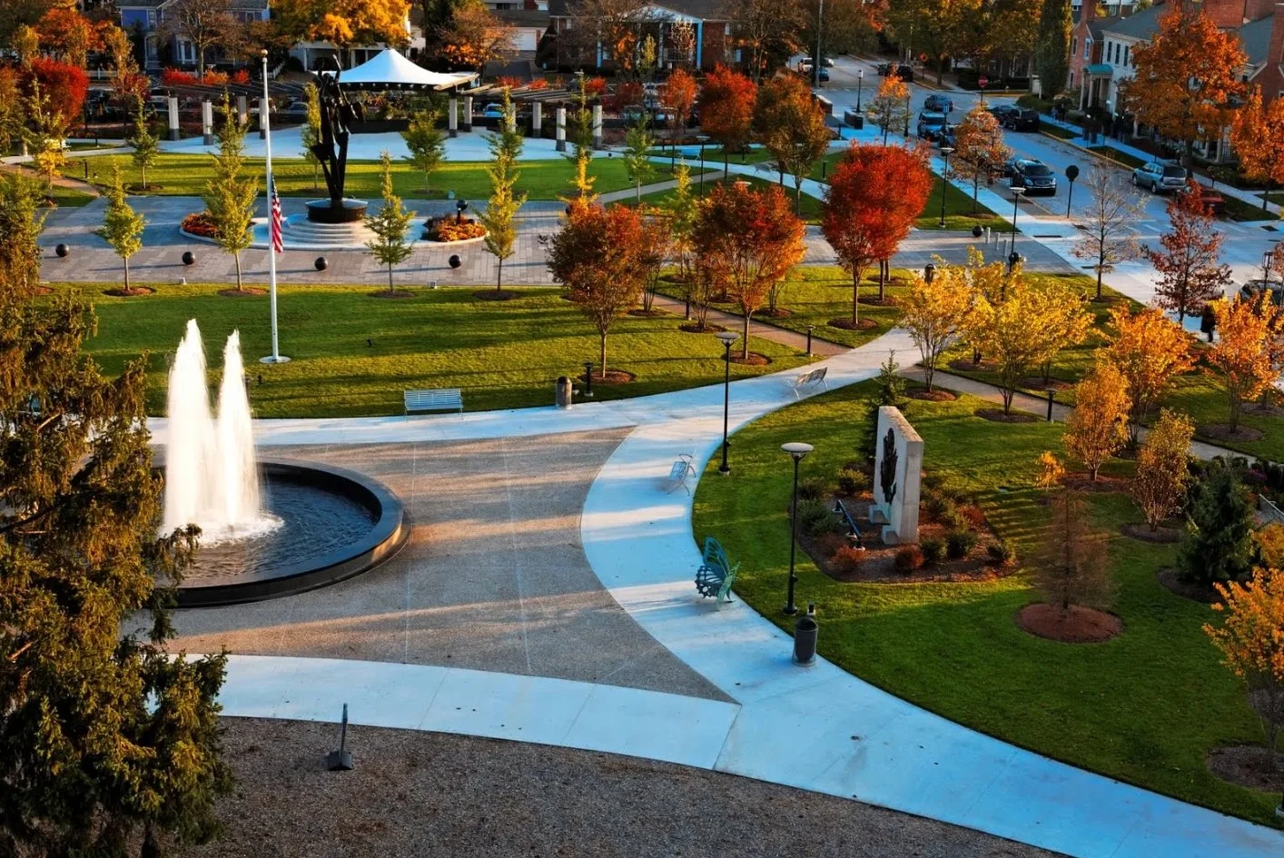 Aerial view of a park with a fountain, trees with autumn leaves, paved walkways, benches, a sculpture, and surrounding buildings and parked cars.