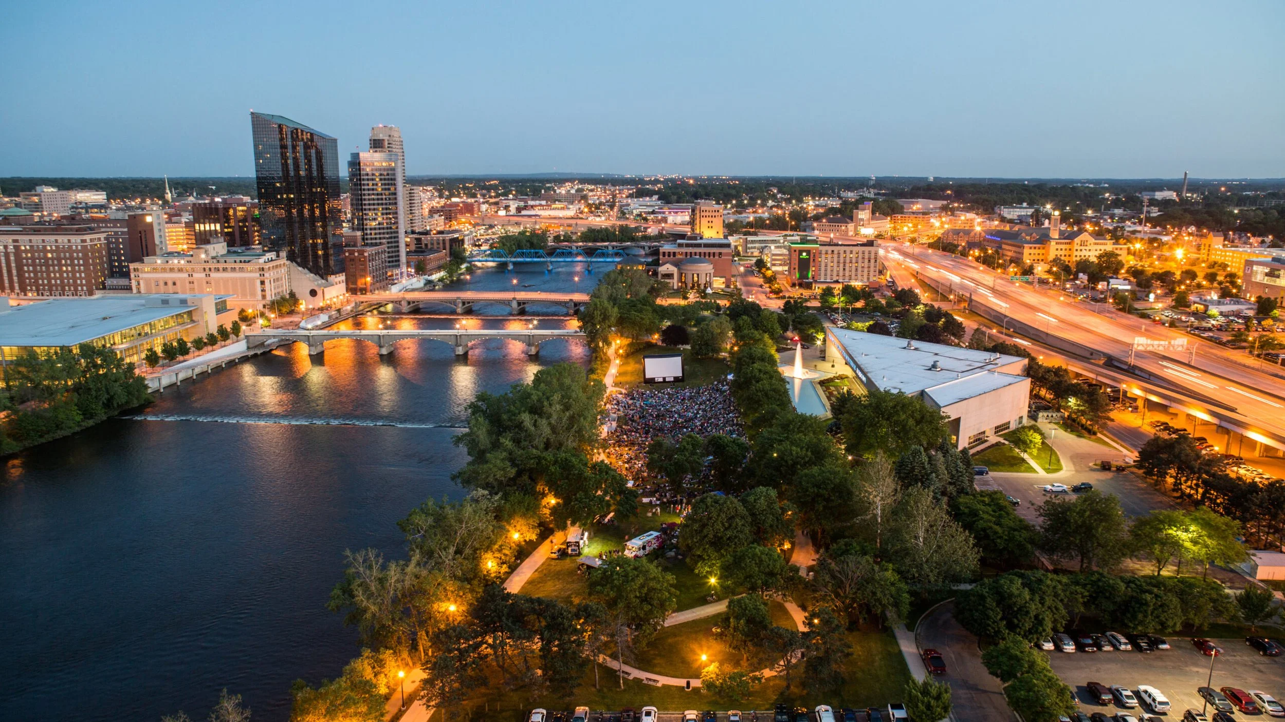 An aerial view of a city skyline at dusk featuring a river with a small waterfall, illuminated bridges, tall buildings, and a park with a crowd gathered near a large outdoor screen.