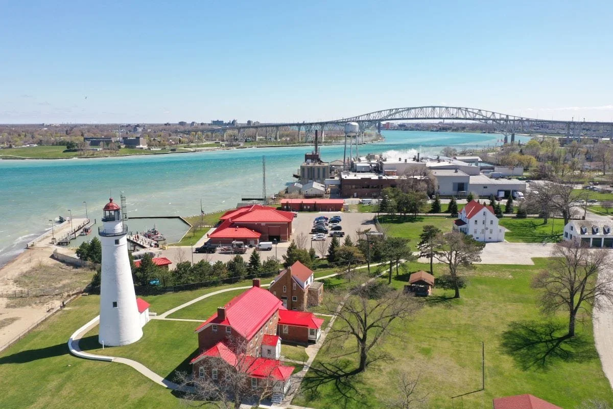 A aerial view of a lighthouse and several buildings with red roofs near a river with a bridge in the background.