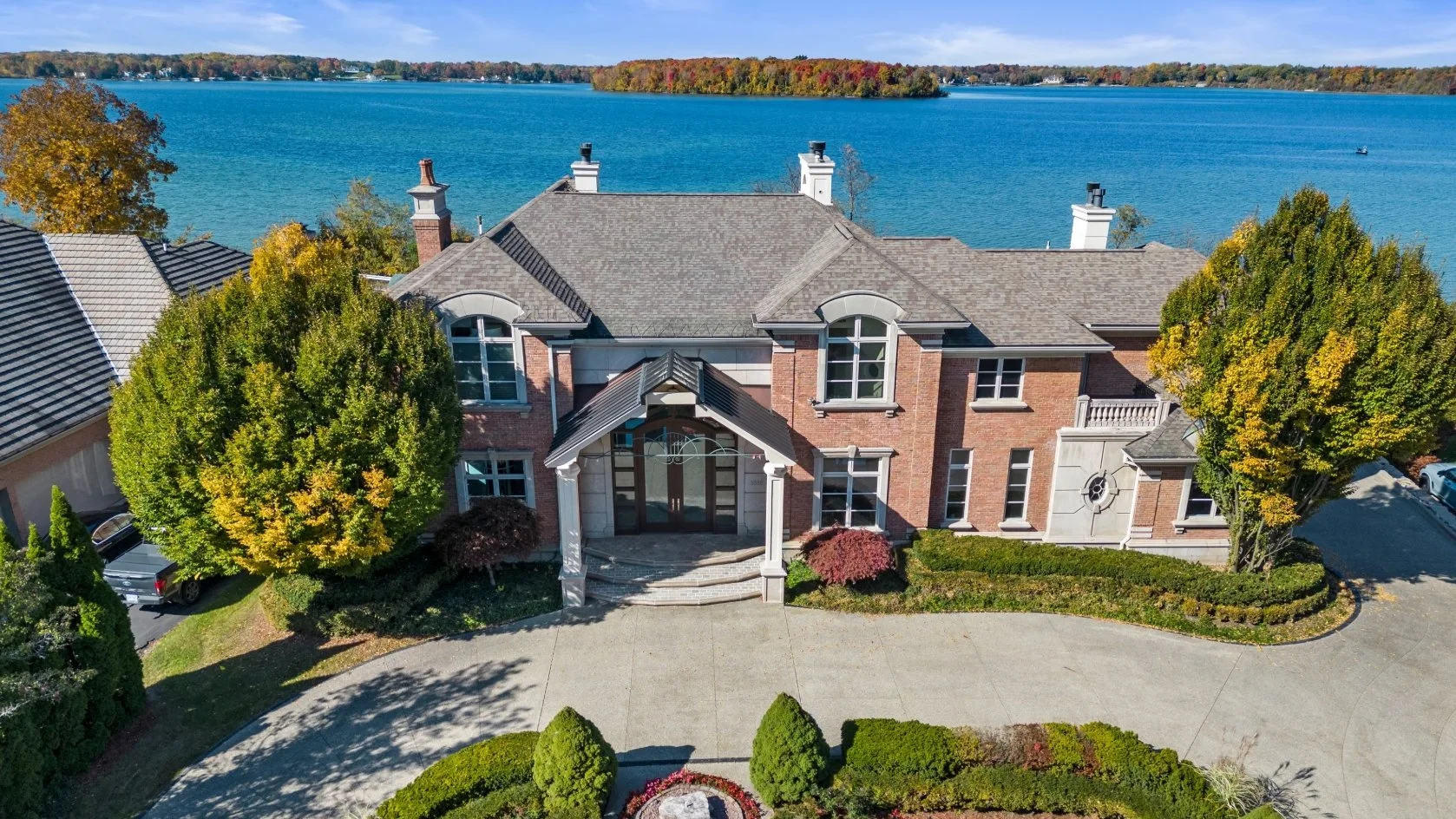 A large brick house with a gray roof and a landscaped front yard by a lake with trees and a boat in the distance.