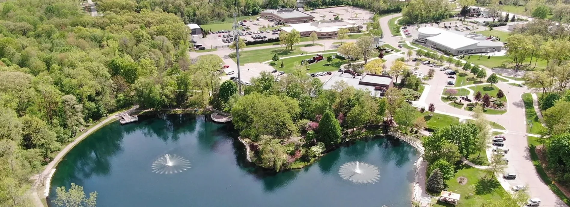 An aerial view of a park with a pond featuring two fountains, surrounded by trees, pathways, and buildings with parking lots.