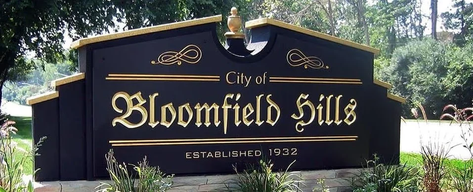 Black sign with gold lettering that reads 'City of Blossomfield Hills, Established 1932', surrounded by plants and trees.