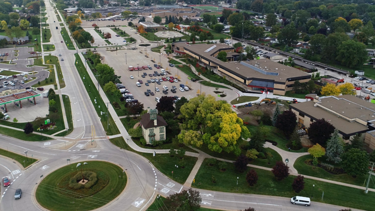 Aerial view of a small town area with a parking lot, roads, green spaces, trees, and buildings, including a shopping center and a gas station.
