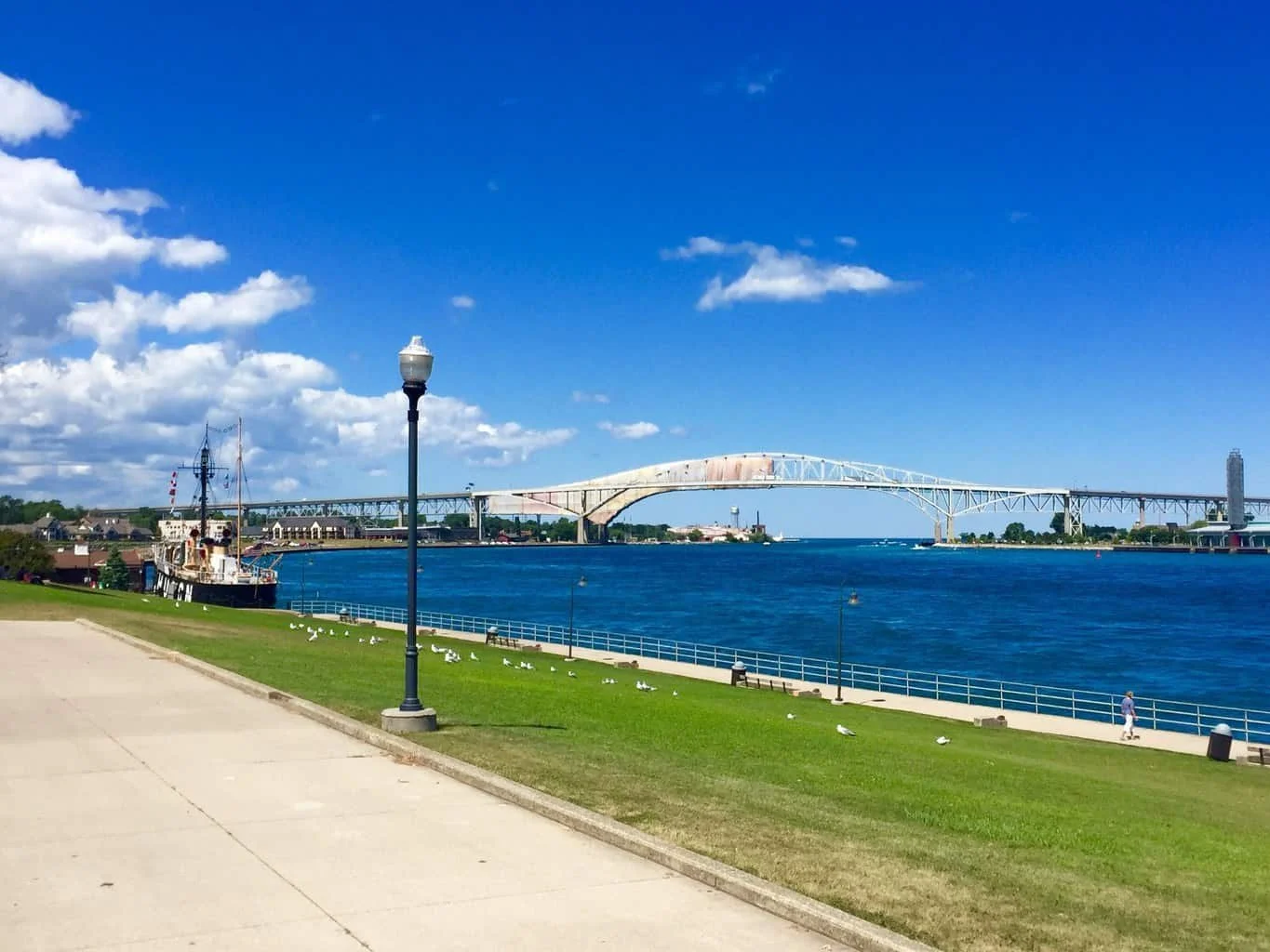 A riverside park with a paved walkway, grassy area, a lamppost, seagulls, and a person walking. In the river, there is a ship and a large bridge in the background under a blue sky with scattered clouds.